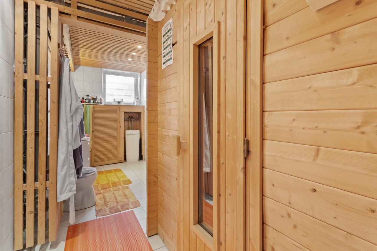 Badezimmer mit Holzsauna, Toilette und Waschbecken, helle Fliesen und bunte Teppiche.