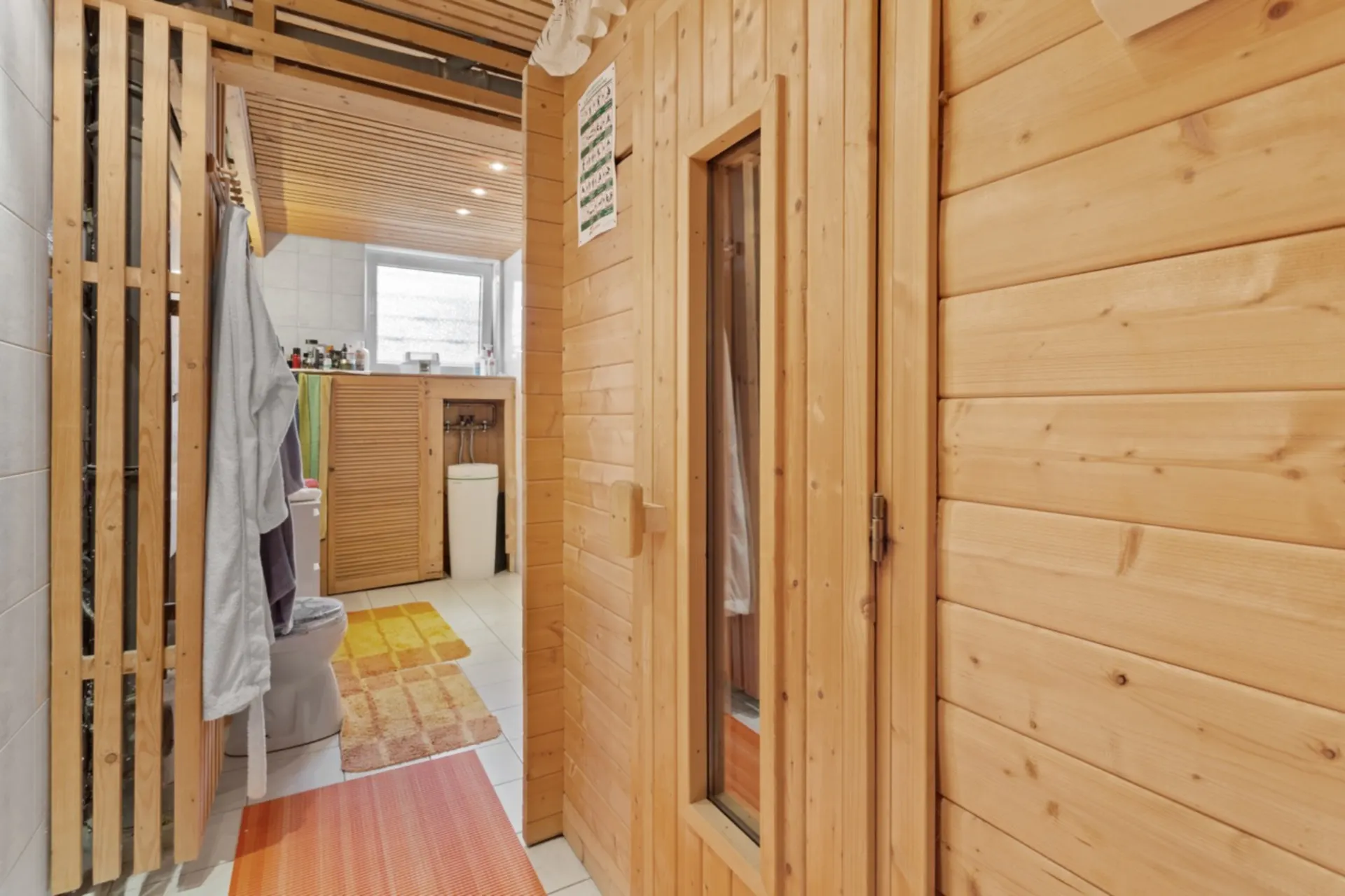 Badezimmer mit Holzsauna, Toilette und Waschbecken, helle Fliesen und bunte Teppiche.