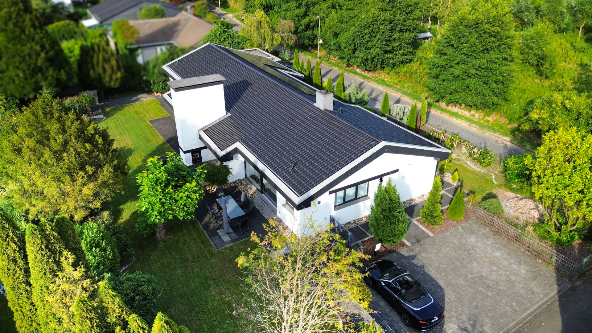 Luftaufnahme eines modernen Hauses mit Solardach, Garten und geparktem Auto in der Einfahrt.