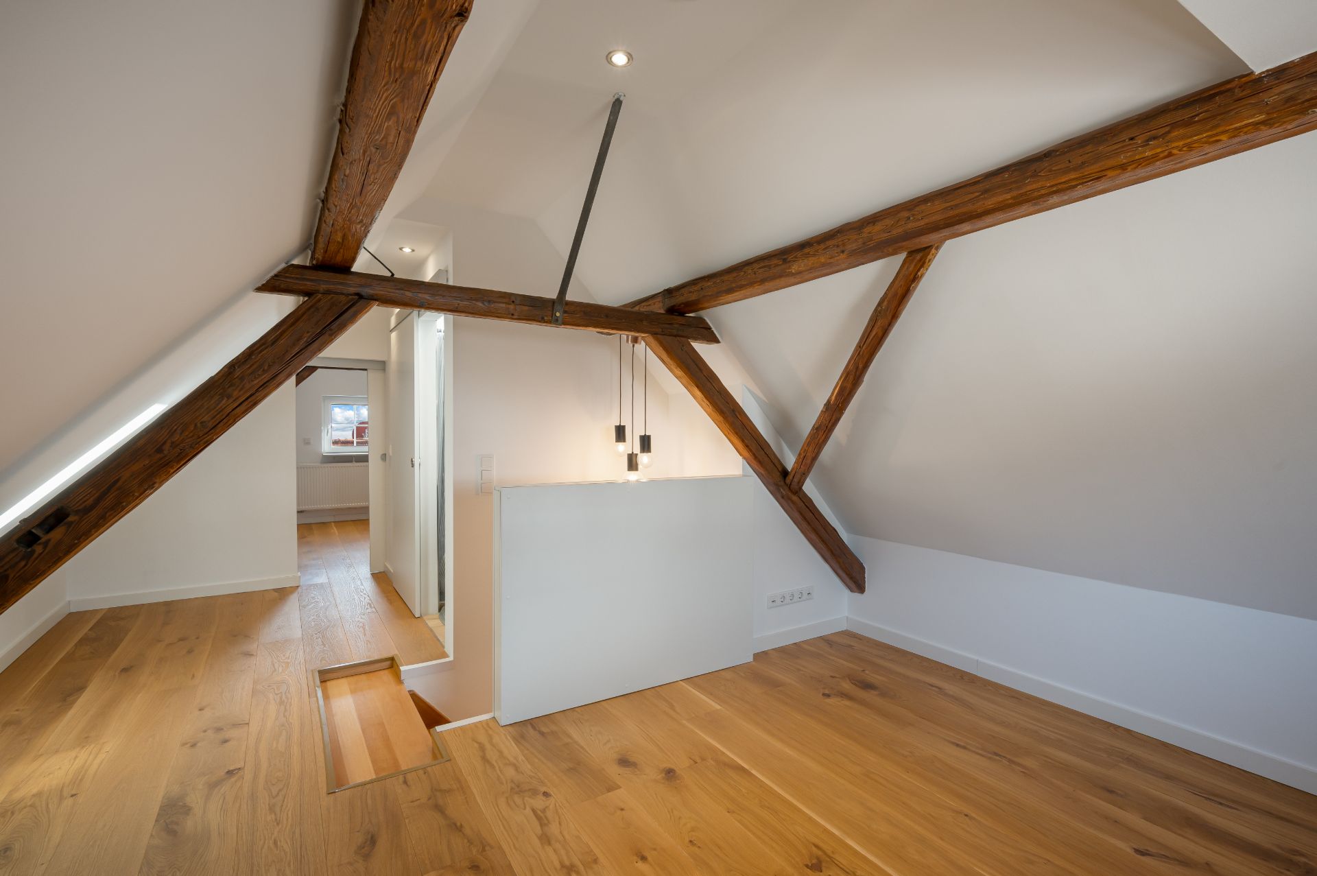Dachzimmer mit sichtbaren Holzbalken, Holzfußboden und weiß gestrichenen Wänden.