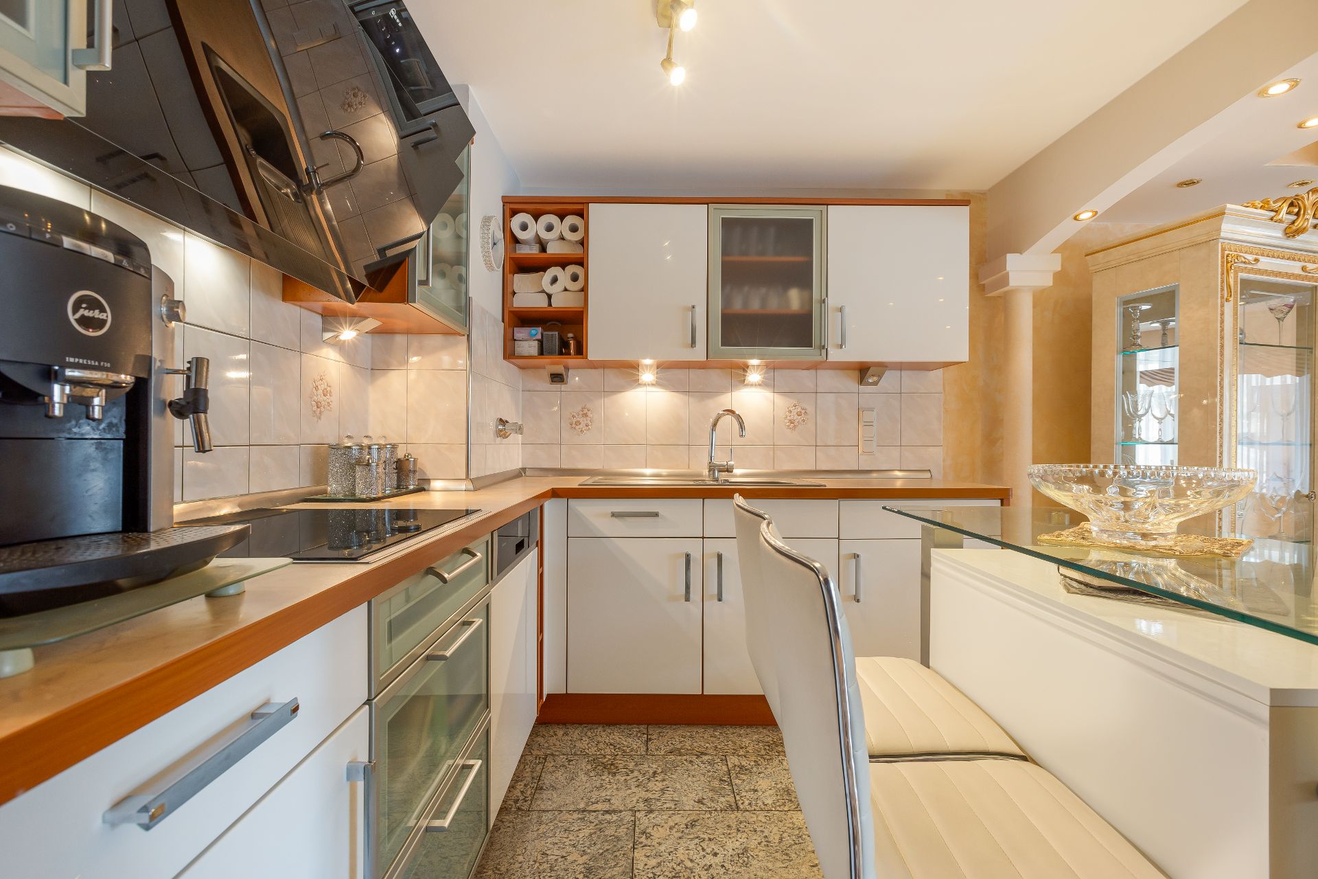 Moderne Küche mit weißen Schränken, Glastisch, Kaffeemaschine und beleuchteter Spüle.