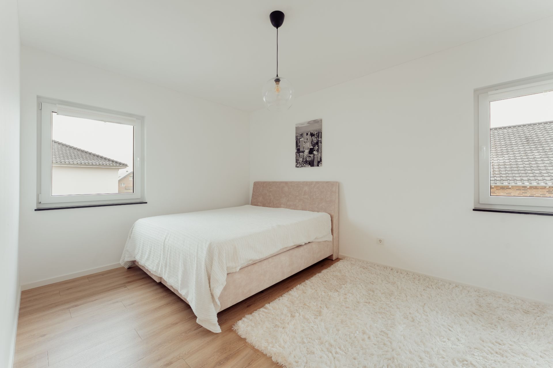 Helles Schlafzimmer mit Doppelbett, zwei Fenstern, Holzfußboden und weißem Teppich.