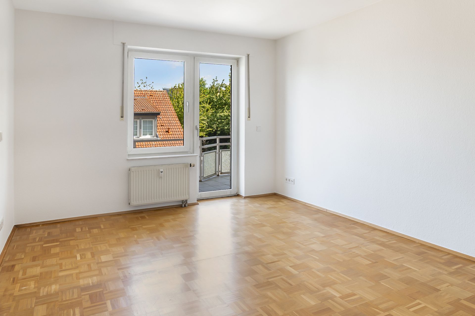 Helles, leeres Zimmer mit Parkettboden, Fenster und Tür zu einem kleinen Balkon.