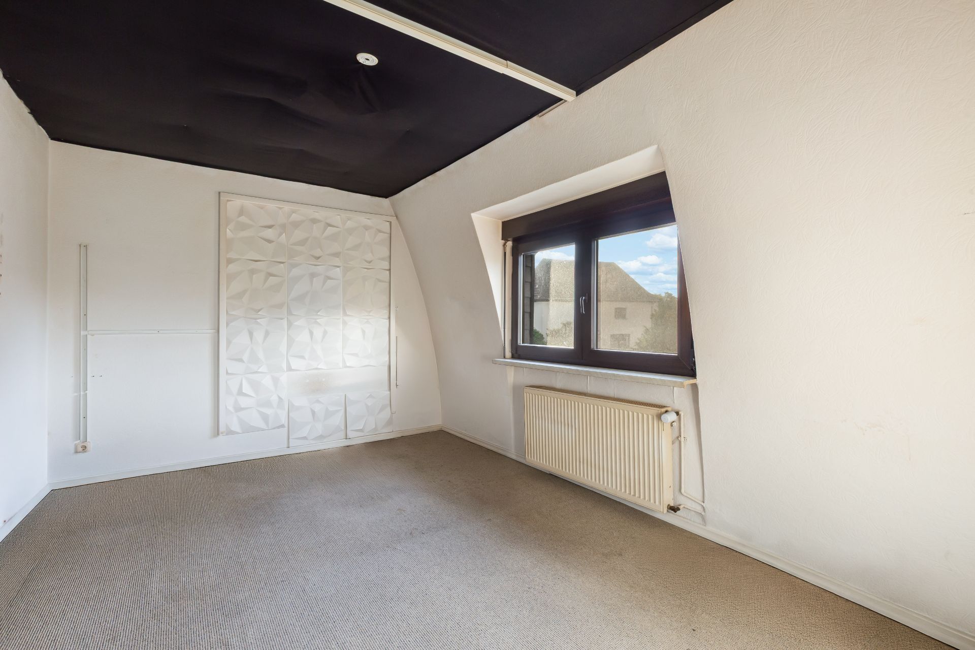 Leeres Zimmer mit Teppichboden, großem Fenster, schwarzer Decke und weißem Heizkörper.