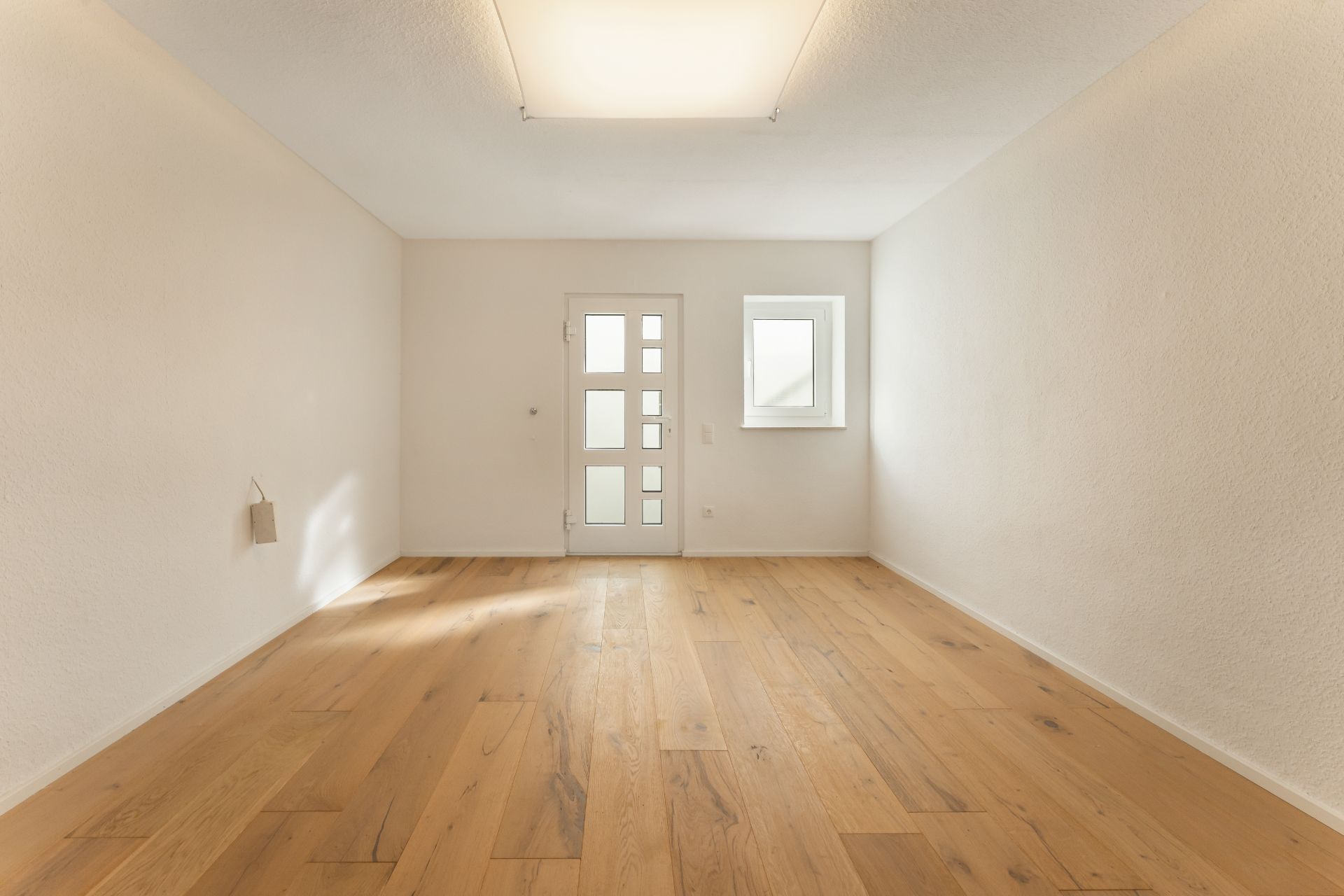 Leerer Raum mit Holzfußboden, weißer Tür und kleinem Fenster an der Rückwand.
