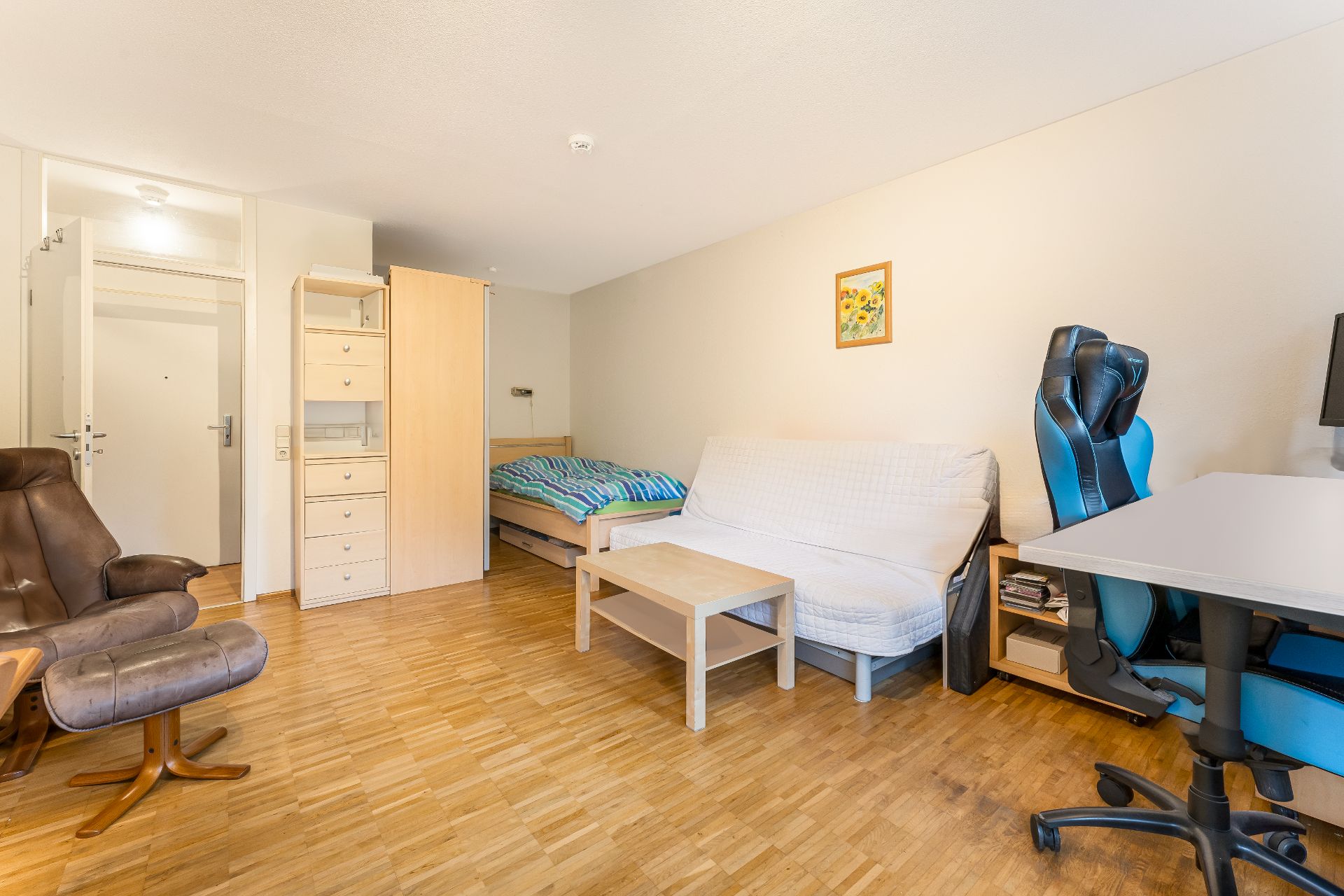 Helles Wohn- und Schlafzimmer mit Sofa, Bett, Schreibtisch und Sessel auf Holzfußboden.