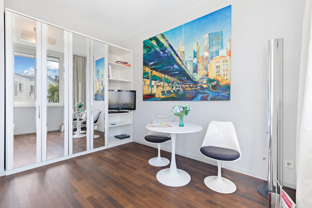 Heller Raum mit weißem Tisch, zwei Stühlen, Spiegelkleiderschrank und buntem Stadtbild an der Wand.