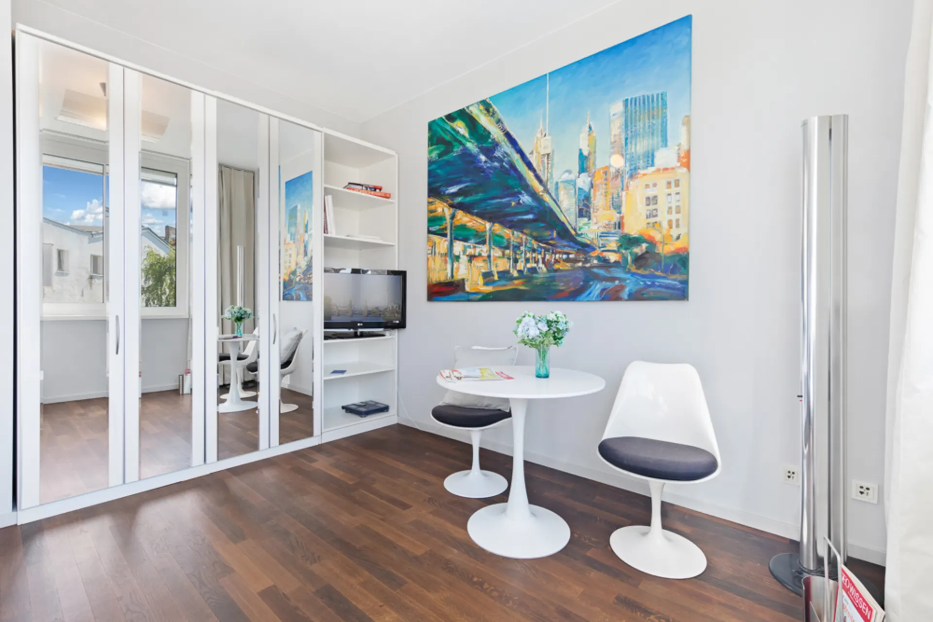 Heller Raum mit weißem Tisch, zwei Stühlen, Spiegelkleiderschrank und buntem Stadtbild an der Wand.