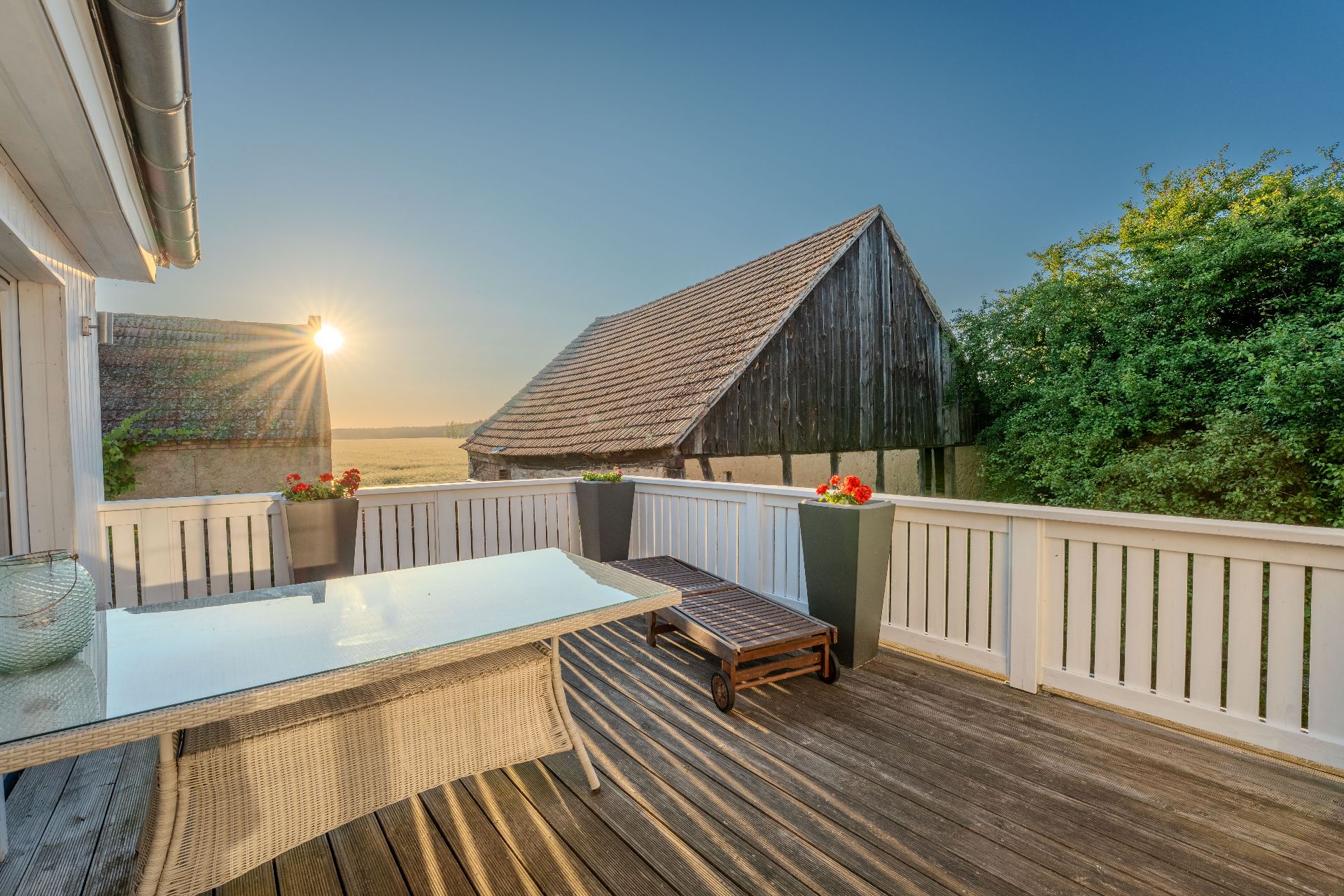 Holzterrasse mit Glastisch, Liege und Blumenkübeln, Blick auf Scheune und Sonnenuntergang.