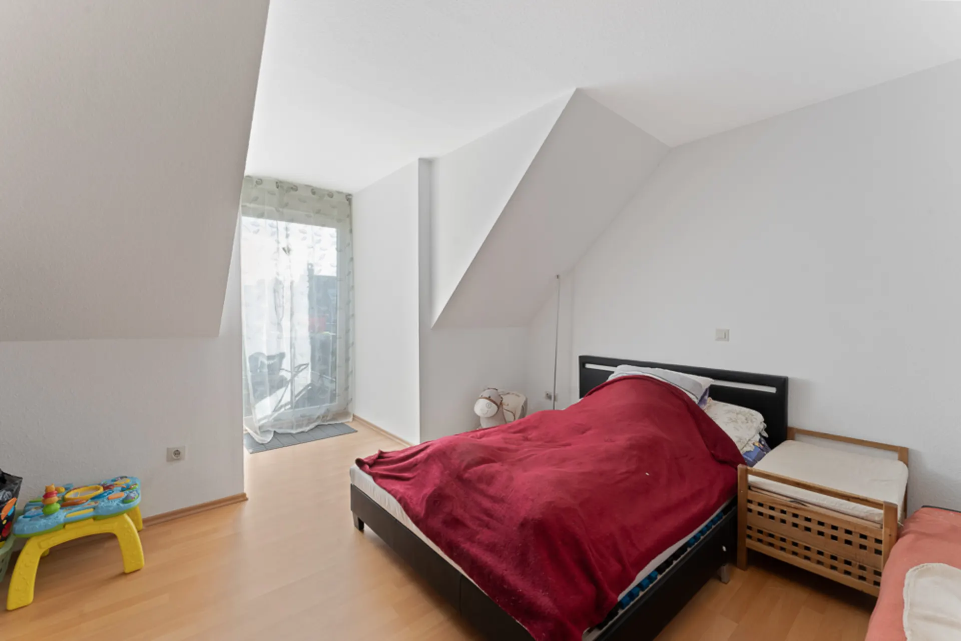 Helles Schlafzimmer mit Doppelbett, Beistellbett und Kinderspieltisch, Laminatboden, Zugang zum Balkon.