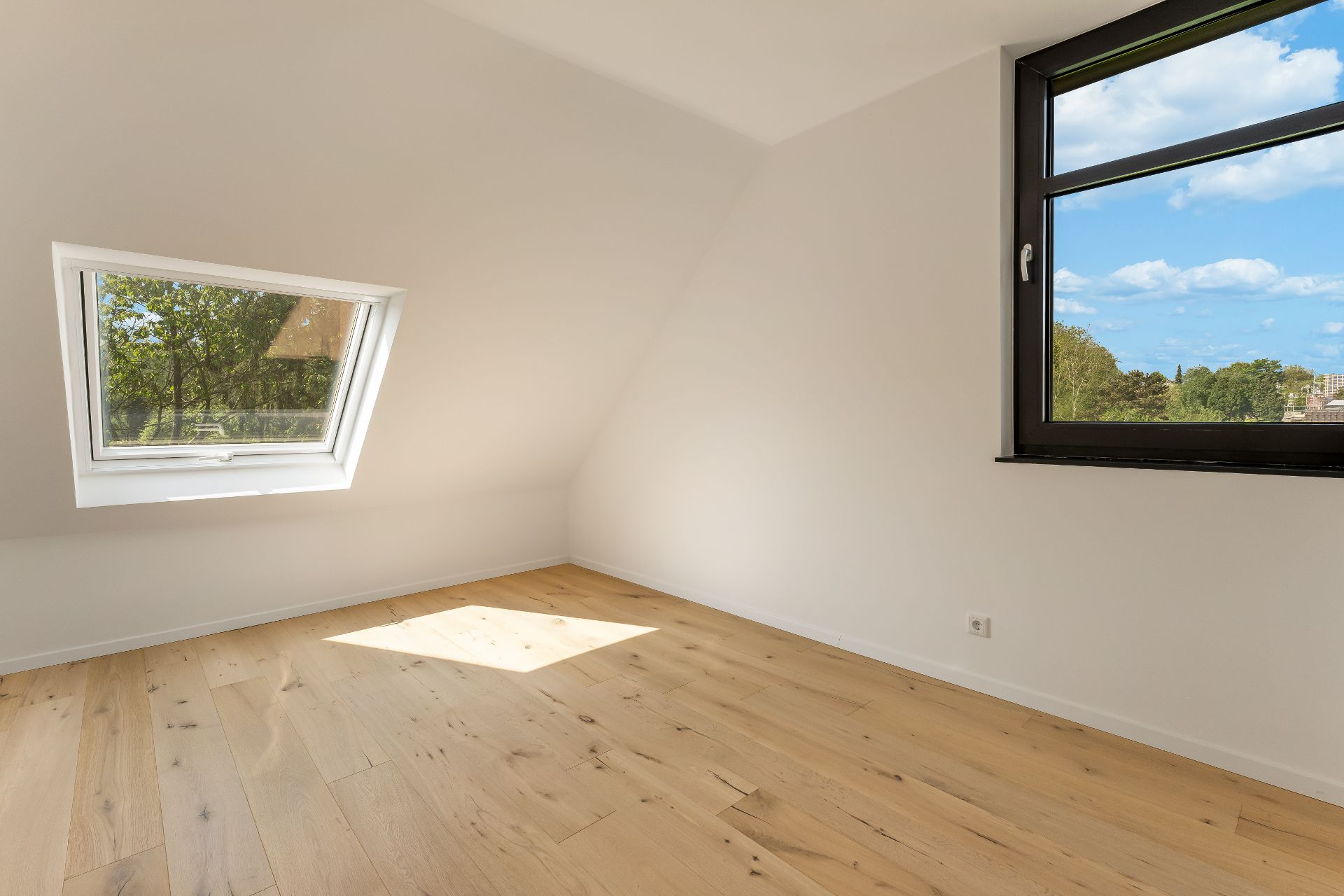 Helles Zimmer mit schrägem Dachfenster, großem Fenster und Holzfußboden.