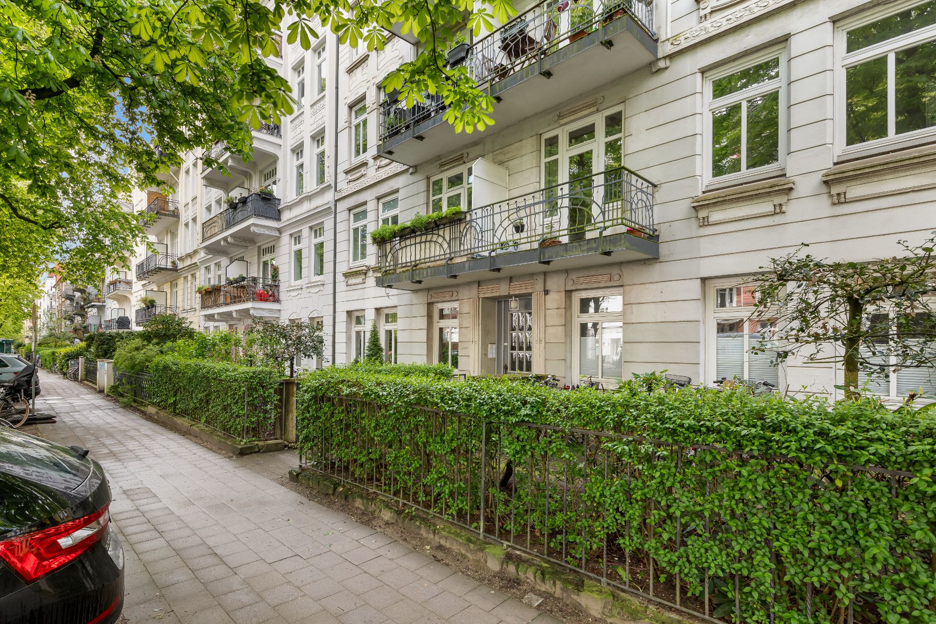 Mehrstöckiges Wohnhaus mit Balkonen, Pflanzen und grüner Hecke an einem Gehweg.