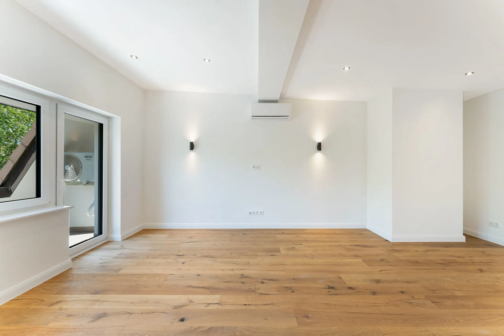 Leerer Raum mit Holzboden, weißen Wänden, Fenstern und einer Glastür nach draußen.