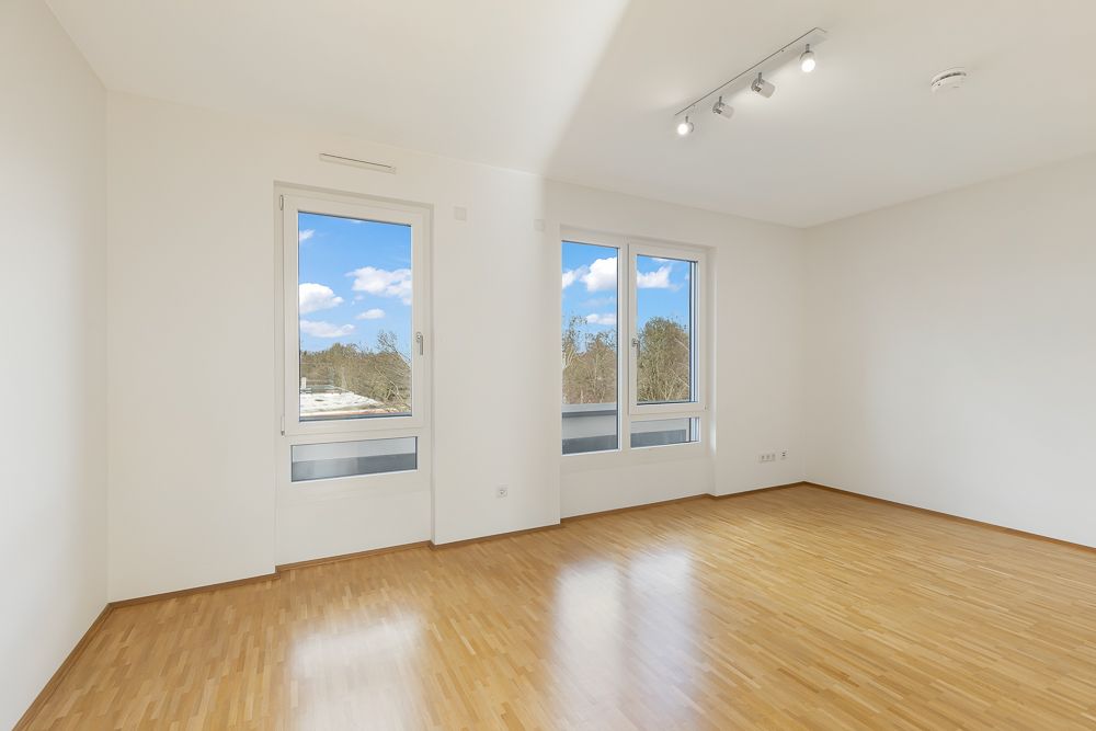 Heller, leerer Raum mit Parkettboden und zwei großen Fenstern, weiße Wände, Deckenstrahler.