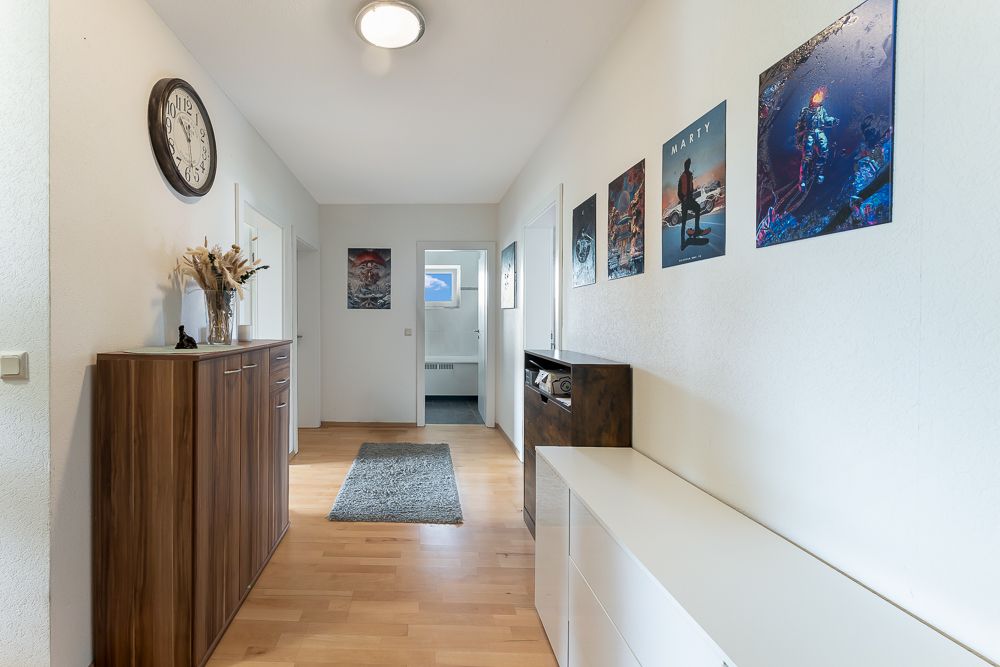 Heller Flur mit Holzboden, Kommode, Wanduhr, Teppich und Bildern an der Wand.