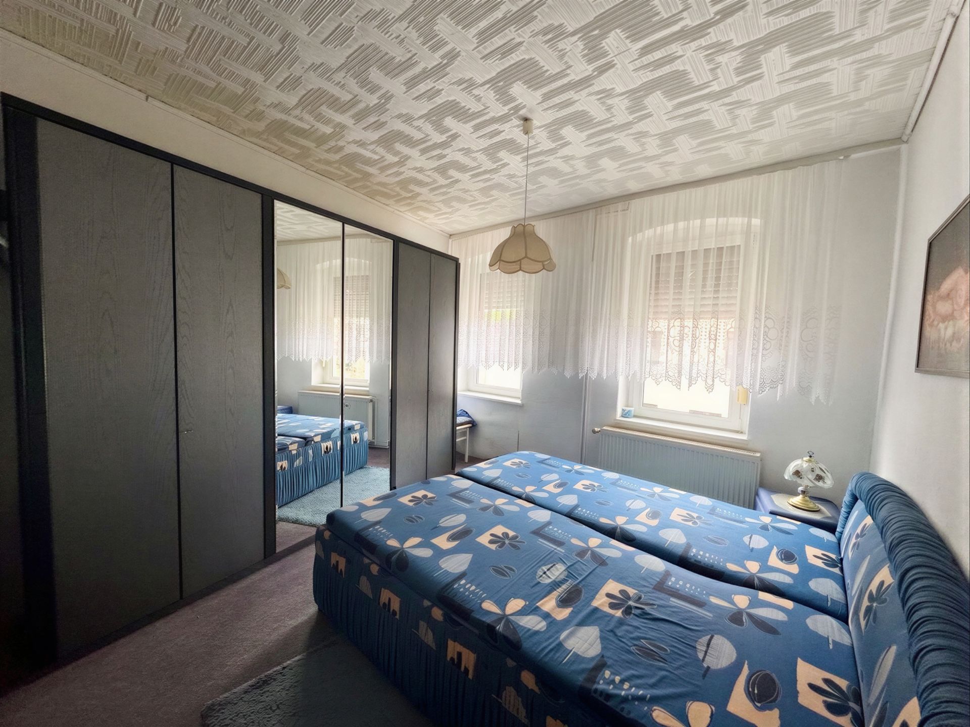 Schlafzimmer mit zwei Betten, großem Kleiderschrank mit Spiegel und zwei Fenstern mit Gardinen.