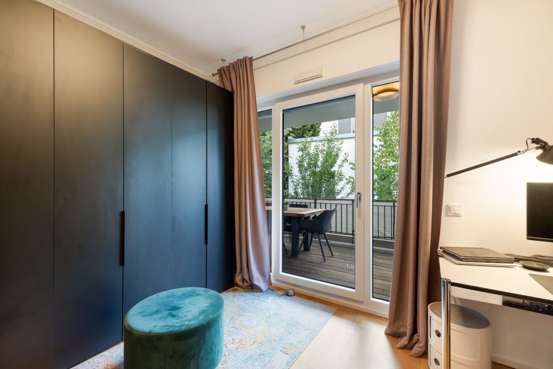 Zimmer mit dunklem Schrank, Schreibtisch, Teppich und Zugang zu einem Balkon mit Tisch und Stühlen.