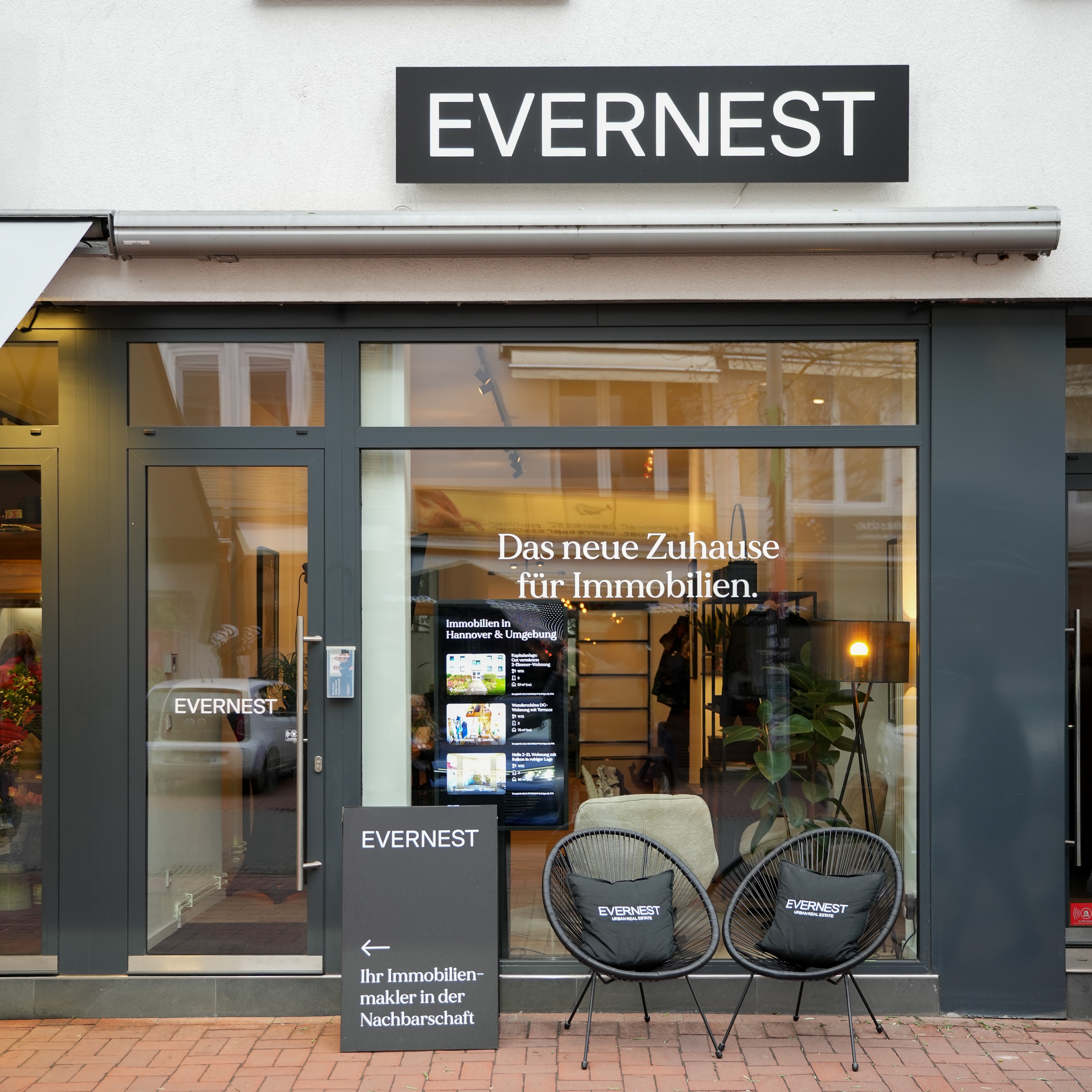 Außenansicht eines Immobilienbüros mit Glasfront, zwei Stühlen und einem Werbeschild vor dem Eingang.