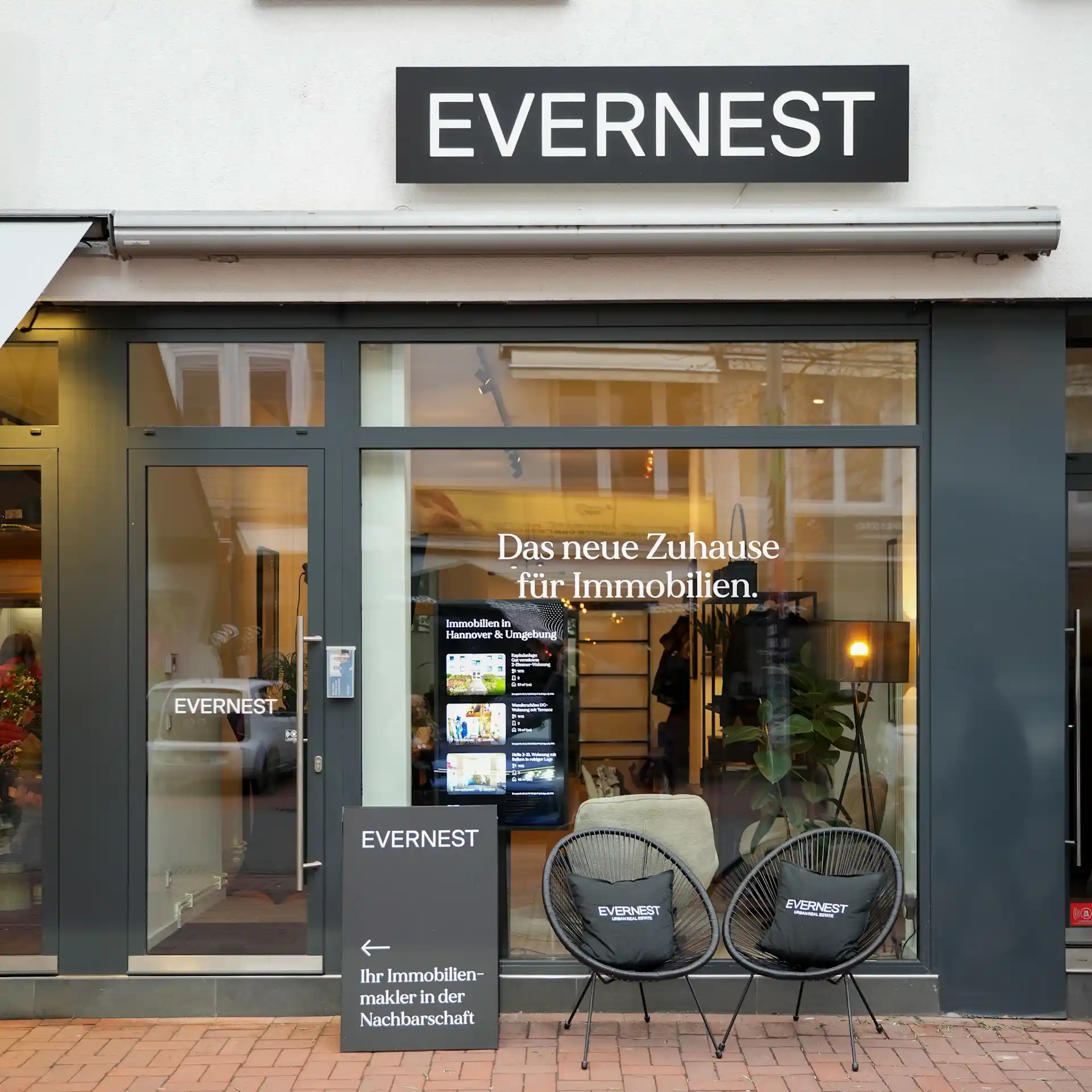 Außenansicht eines Immobilienbüros mit Glasfront, zwei Stühlen und einem Werbeschild vor dem Eingang.