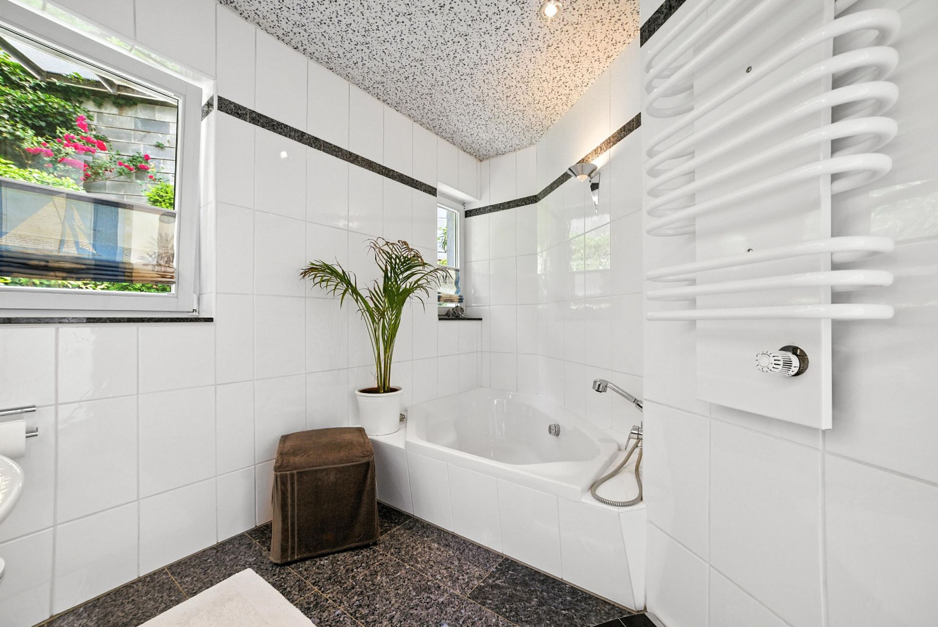 Helles Badezimmer mit Eckbadewanne, weiß gefliesten Wänden, Fenster und einer Pflanze.
