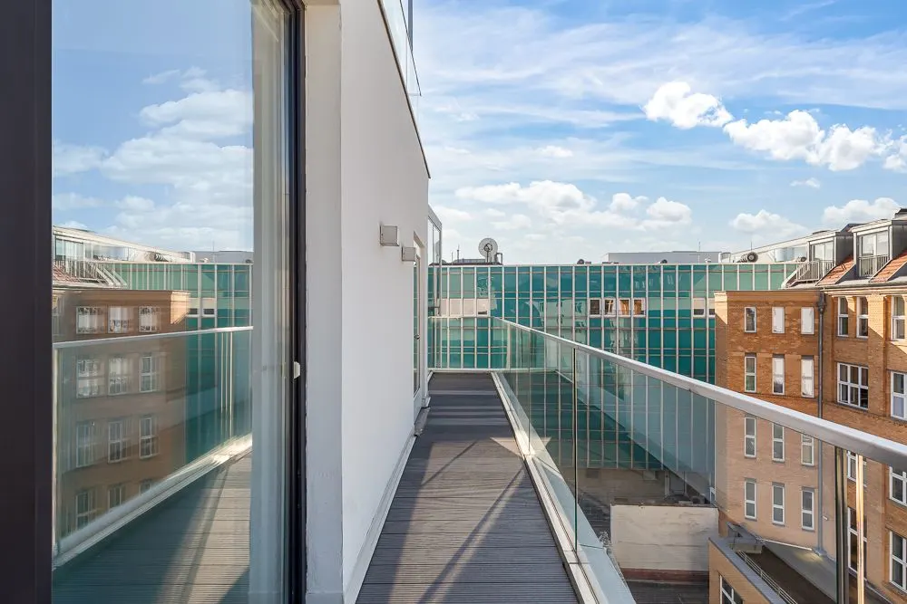 Moderner Balkon mit Glasgeländer, umgeben von Gebäuden, Blick auf blauen Himmel und Wolken.