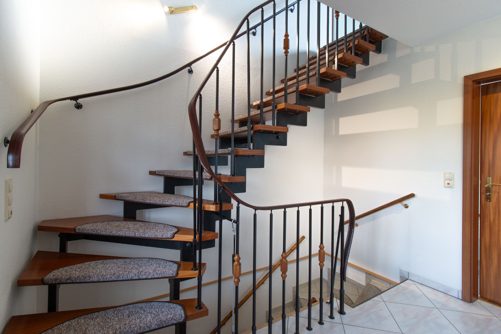 Holztreppe mit Teppichstufen und schwarzem Geländer in einem Flur mit weißer Wand.