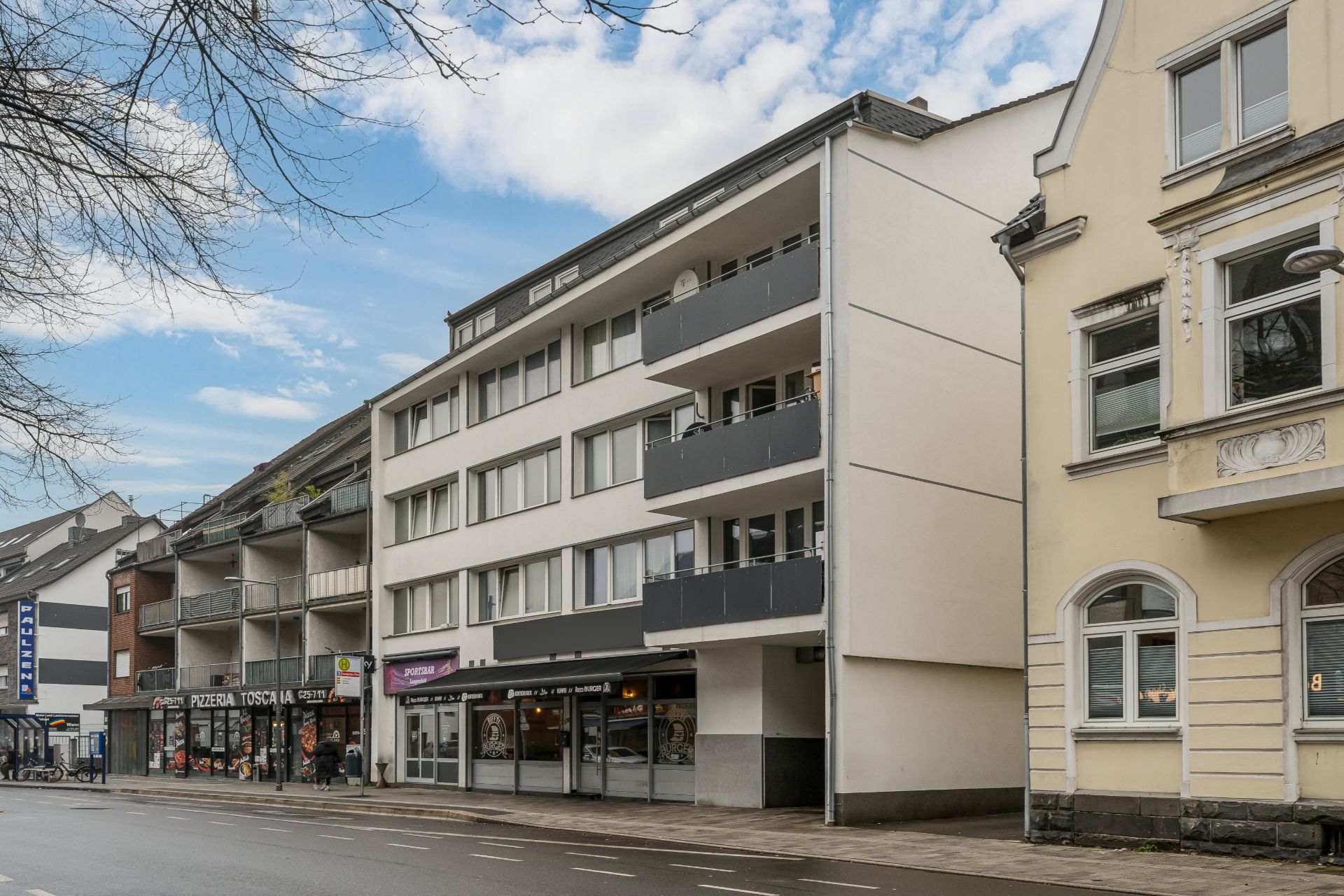 Mehrstöckiges Wohnhaus mit Balkonen und Geschäften im Erdgeschoss an einer Straße.