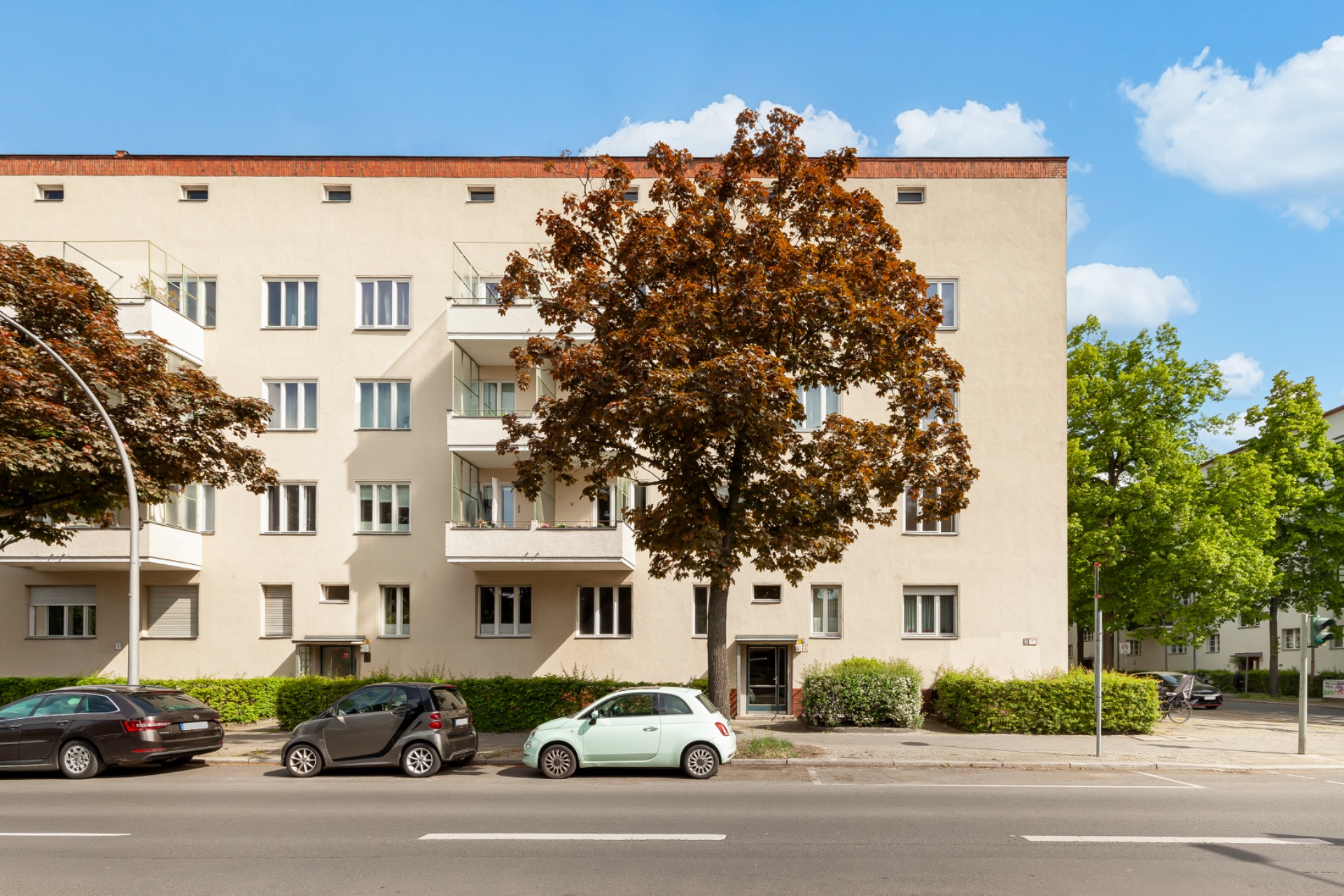 Vierstöckiges Wohnhaus mit Balkonen, Bäumen davor und geparkten Autos an der Straße.