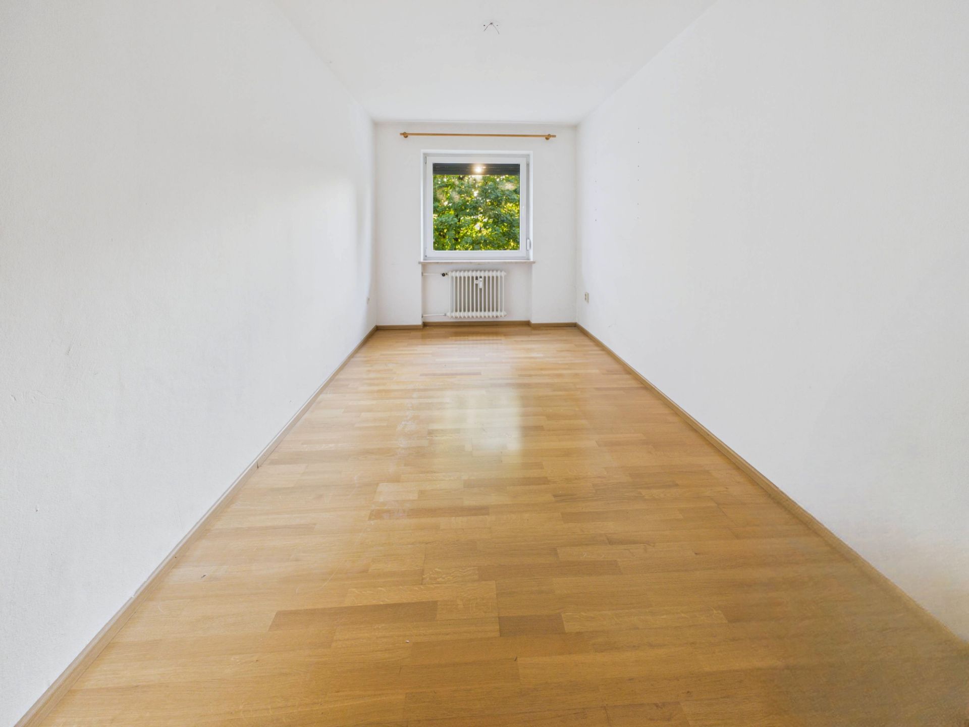 Leerer Raum mit Holzboden, weißen Wänden und Fenster mit Blick ins Grüne.