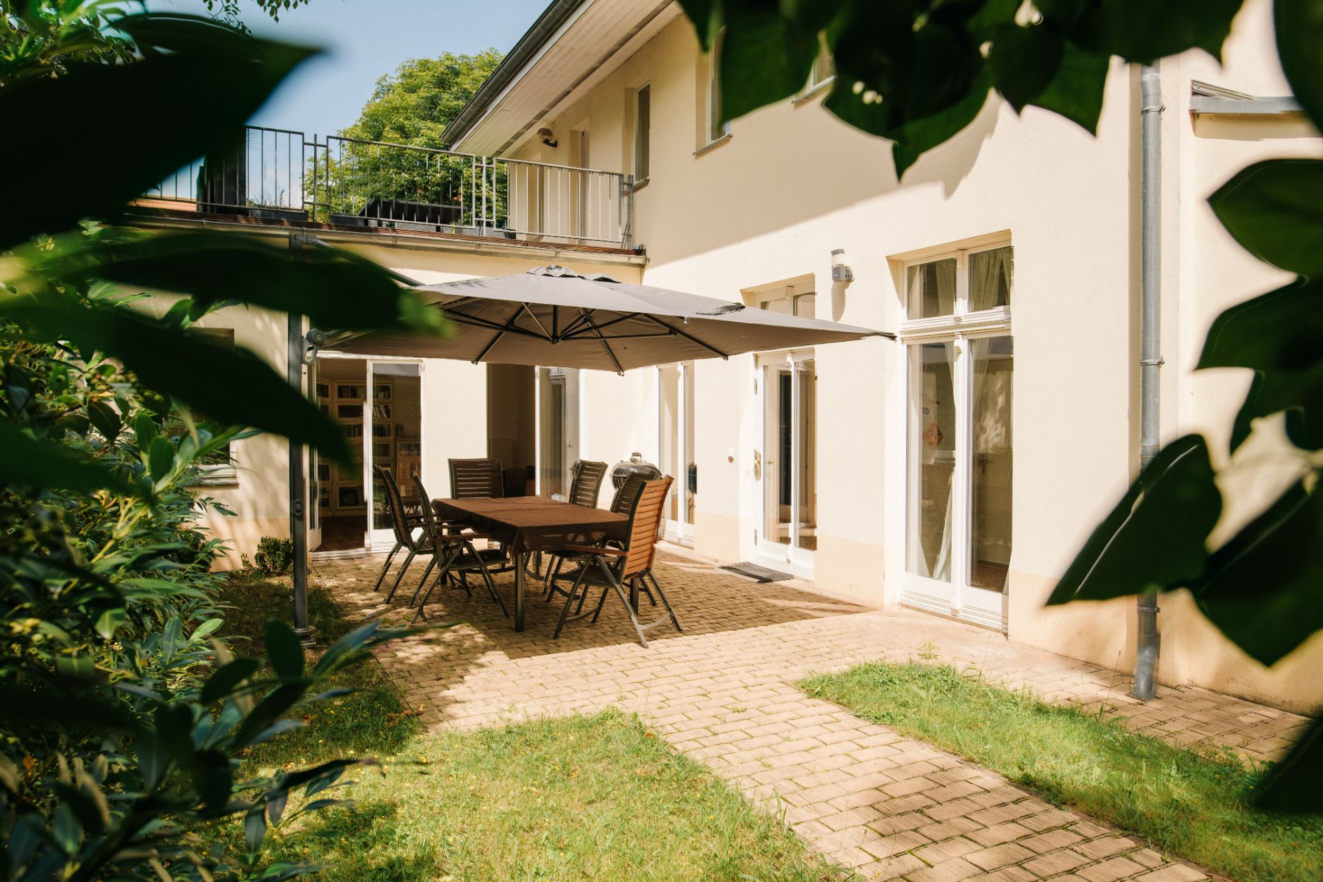 Terrasse mit Tisch, Stühlen und großem Sonnenschirm neben einem Haus, umgeben von Grün.