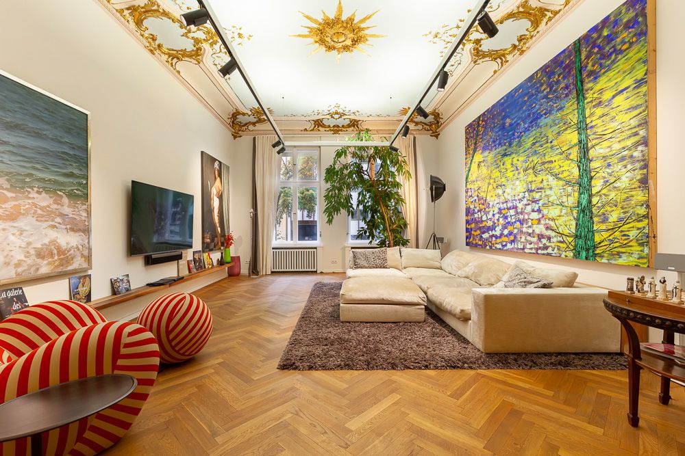 Großes Wohnzimmer mit beigem Sofa, Gemälden, Holzboden und verziertem Deckengemälde.