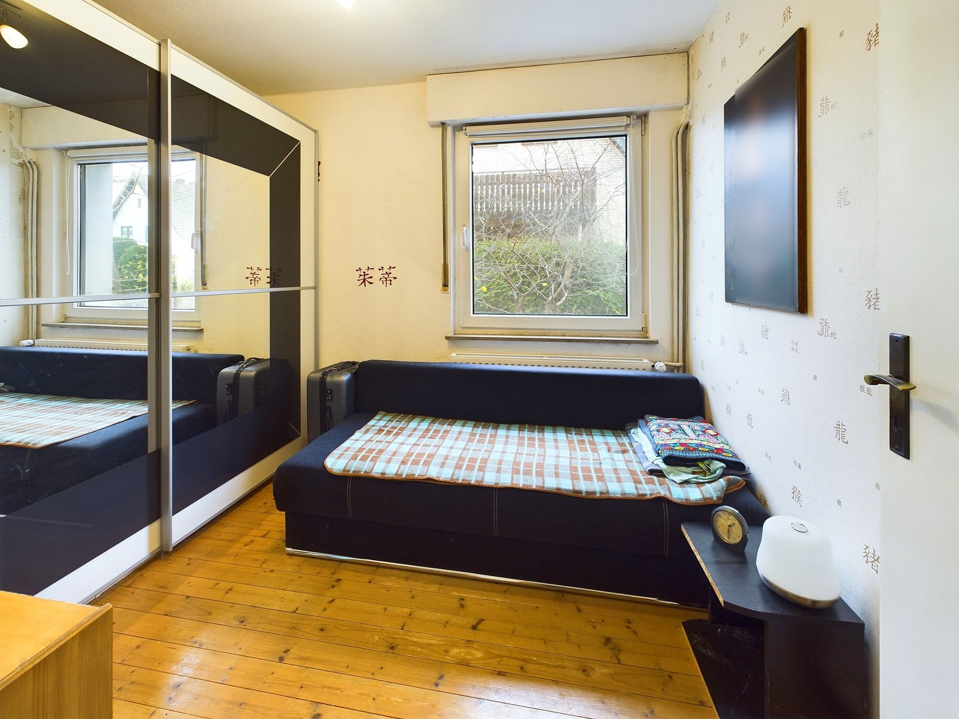 Kleines Zimmer mit Bett, Kleiderschrank mit Spiegel und Fenster, Holzfußboden.