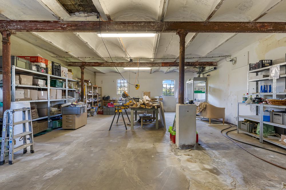 Großer Werkraum mit Regalen, Werkzeugen und Tischen, alte Decke mit sichtbaren Balken.