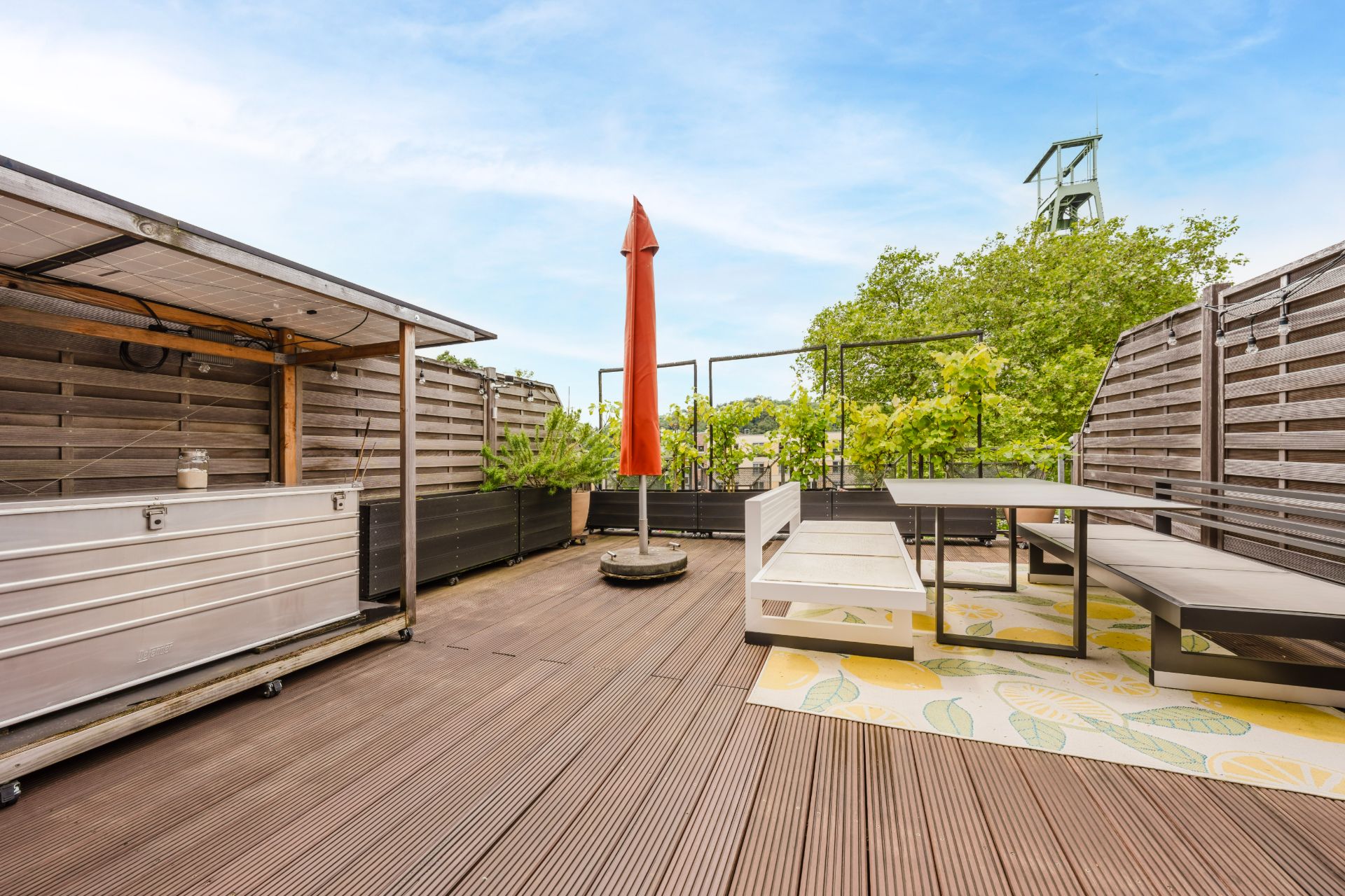 Große Dachterrasse mit Holzboden, Sitzbank, Tisch und rotem Sonnenschirm, umgeben von Holzzäunen.