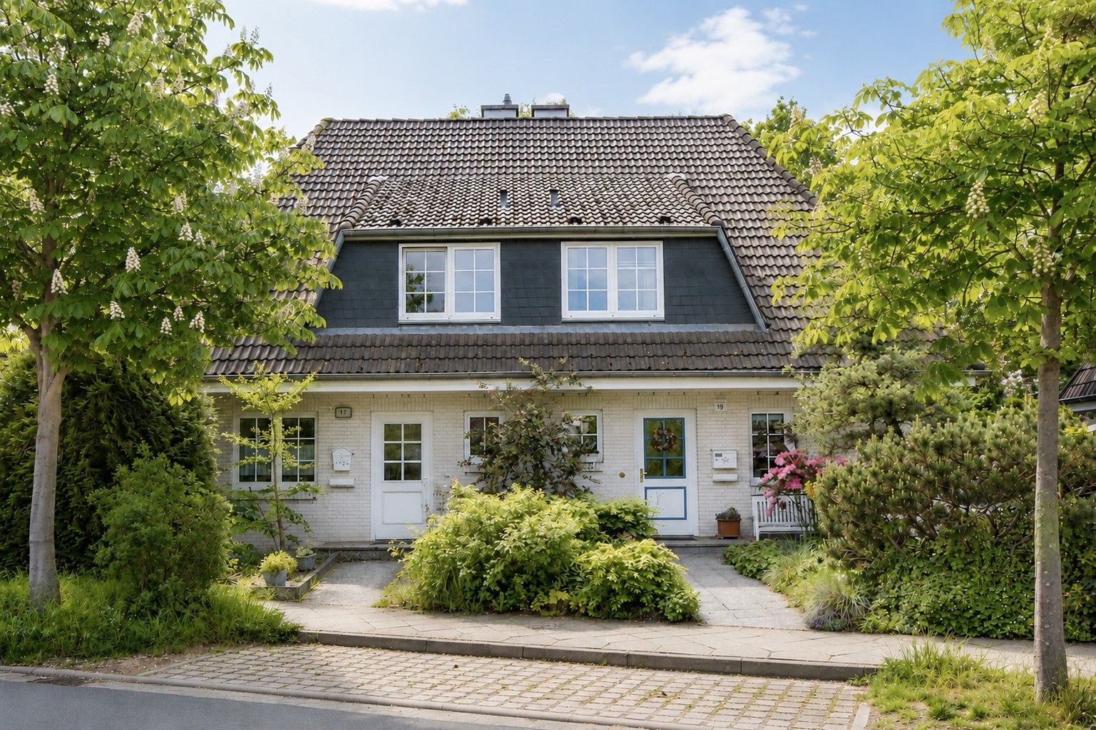 Doppelhaushälfte mit großem Dach und Vorgarten, umgeben von Bäumen und Sträuchern.
