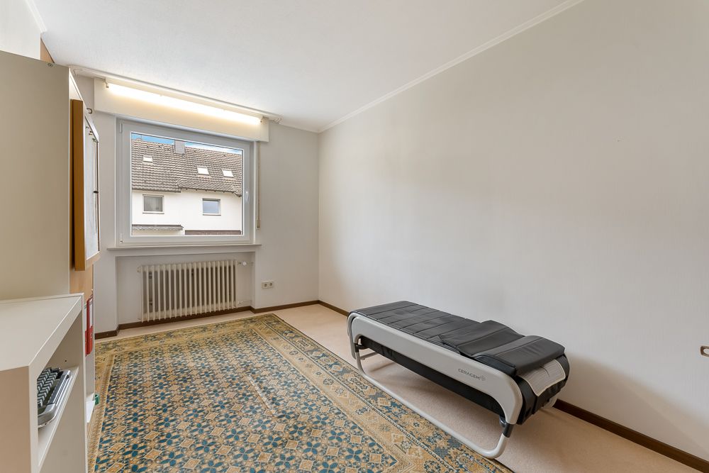 Helles Zimmer mit Teppich, Fenster, Heizung und einer Liege am rechten Rand.
