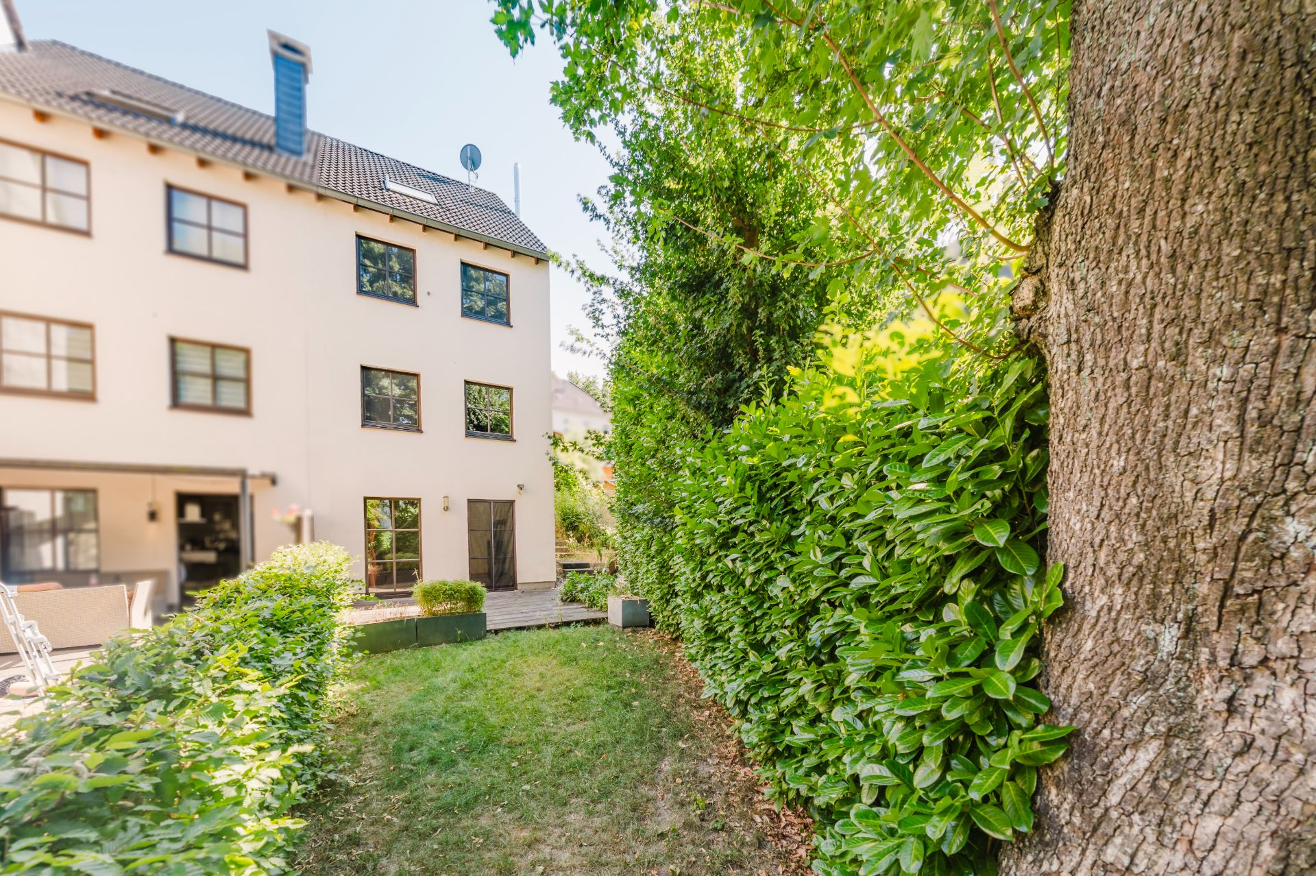 Garten mit Rasenfläche, Hecke und Baum, im Hintergrund ein mehrstöckiges Wohnhaus.