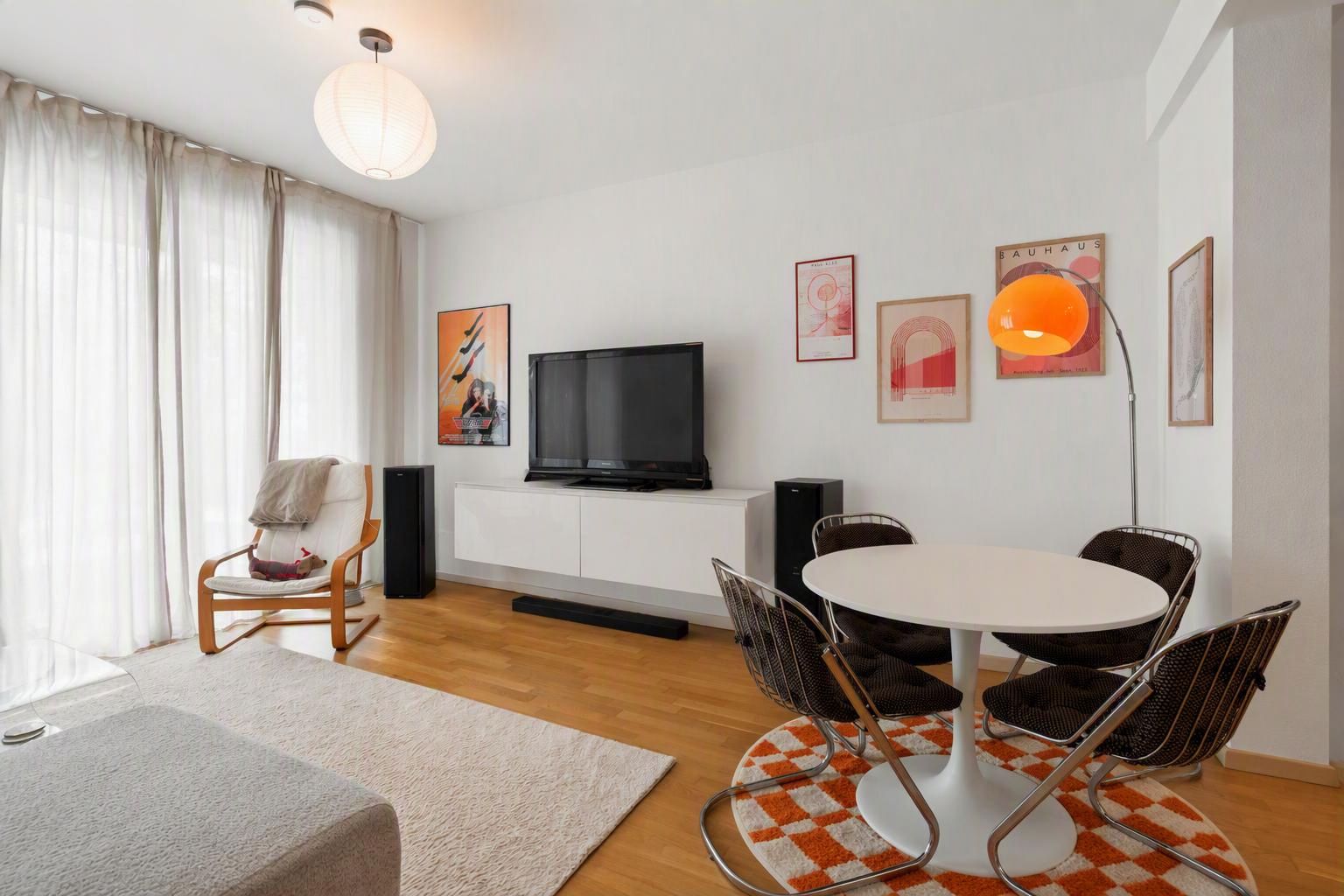 Modernes Wohnzimmer mit Fernseher, Esstisch, vier Stühlen und großer Fensterfront mit Vorhängen.