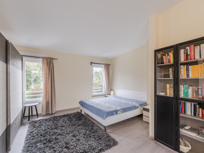 Schlafzimmer mit Doppelbett, Bücherregal, Teppich und zwei Fenstern mit Vorhängen.