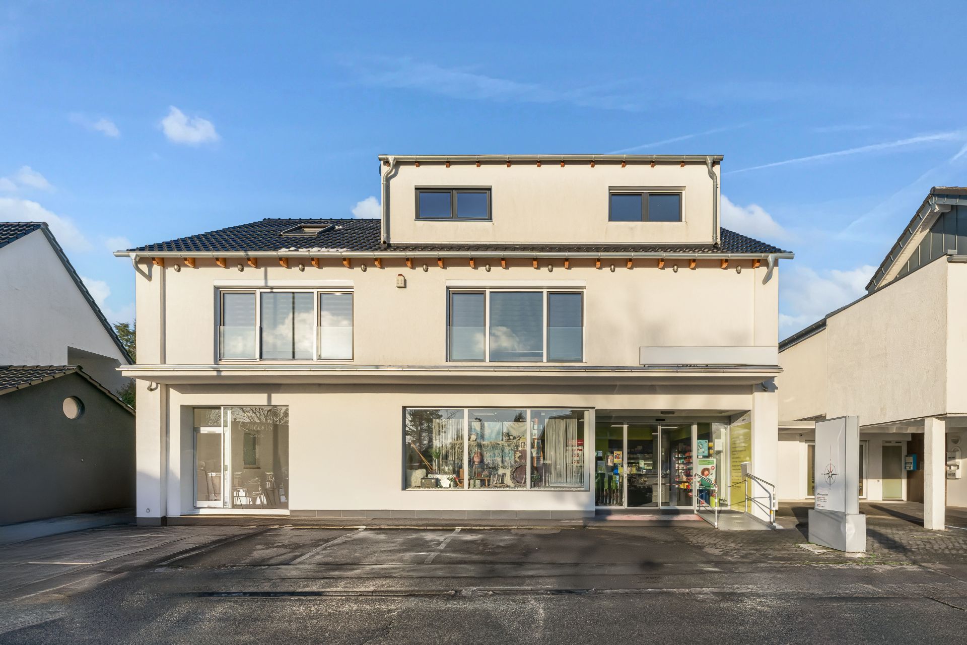 Modernes, weißes Wohngebäude mit großen Fenstern und flachem Dach, davor ein Parkplatz.