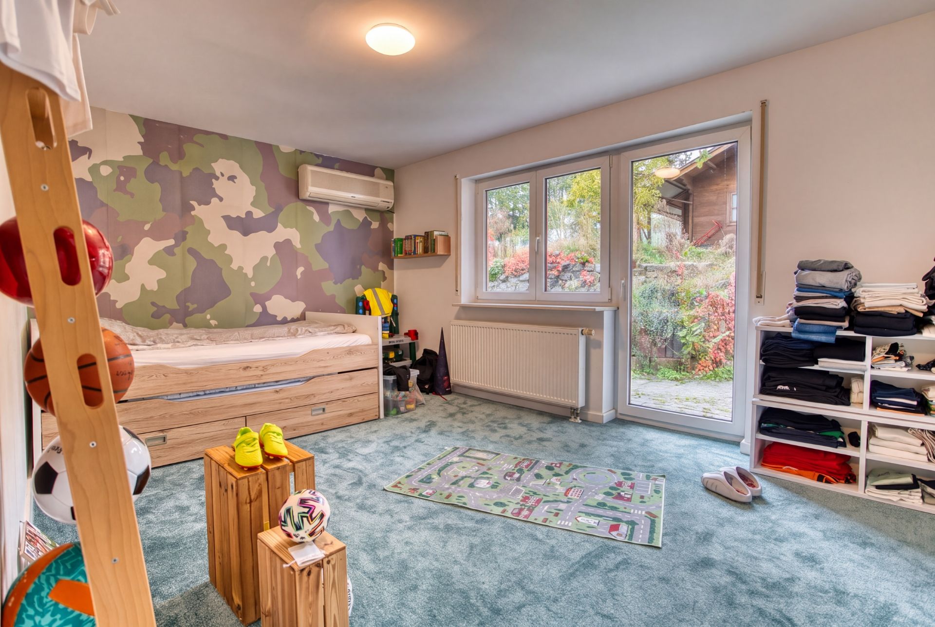 Kinderzimmer mit Einzelbett, grünem Teppich, Wandregal und Ausblick in den Garten.