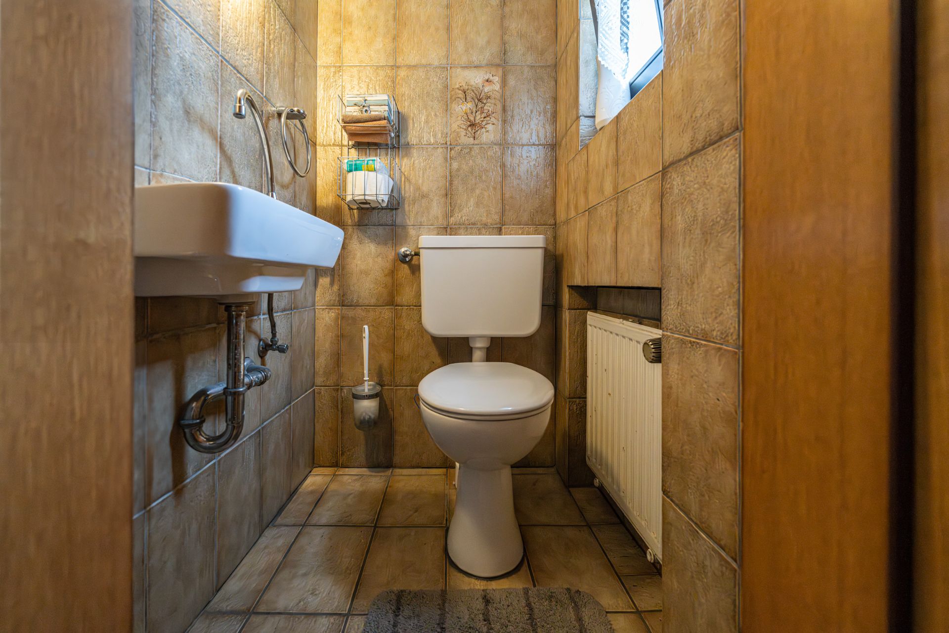 Kleines Bad mit WC, Waschbecken, Heizung und kleinem Fenster, beige gefliest.