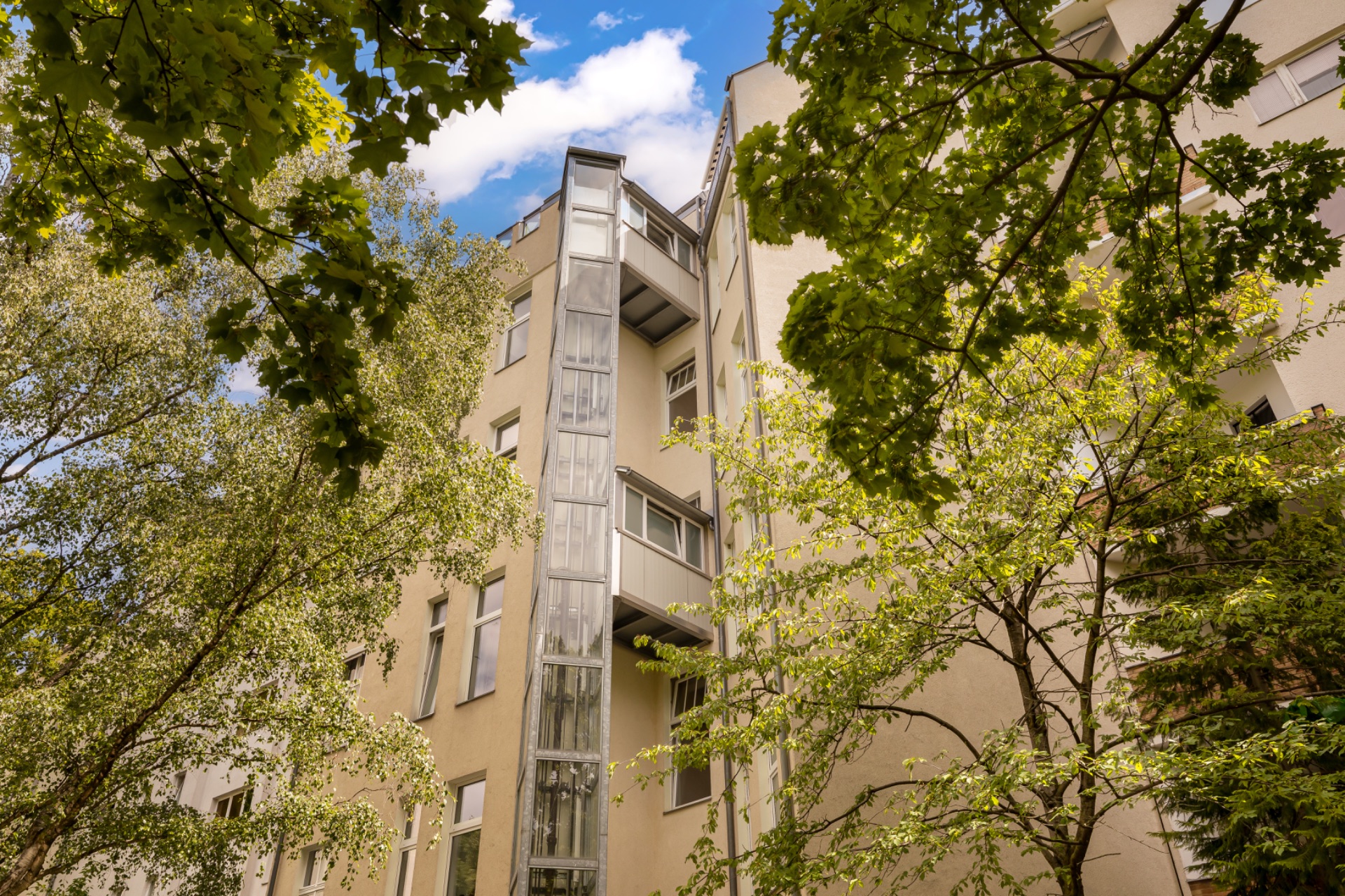 Mehrstöckiges Wohnhaus mit verglastem Aufzug, umgeben von grünen Bäumen.