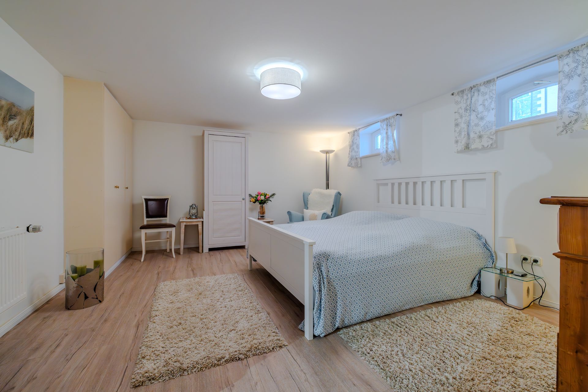Helles Schlafzimmer mit Doppelbett, Teppichen, Sessel, Schrank und zwei kleinen Fenstern.