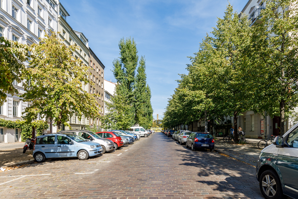 Baumbestandene Wohnstraße mit parkenden Autos und Wohnungen auf beiden Seiten bei Sonnenschein.