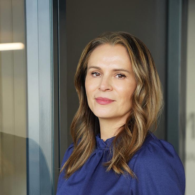 Frau mit langen braunen Haaren und blauem Oberteil steht vor einem Fenster.