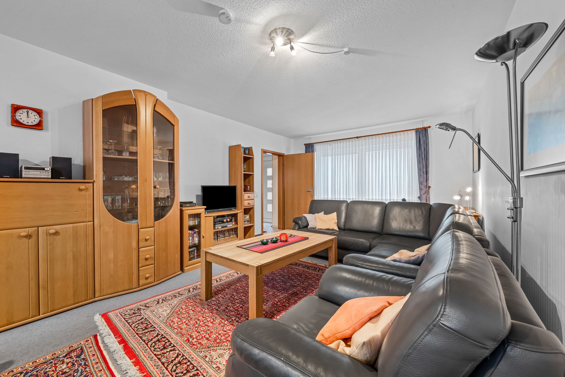 Wohnzimmer mit schwarzem Ledersofa, Holzmöbeln, Teppich und großem Fenster.