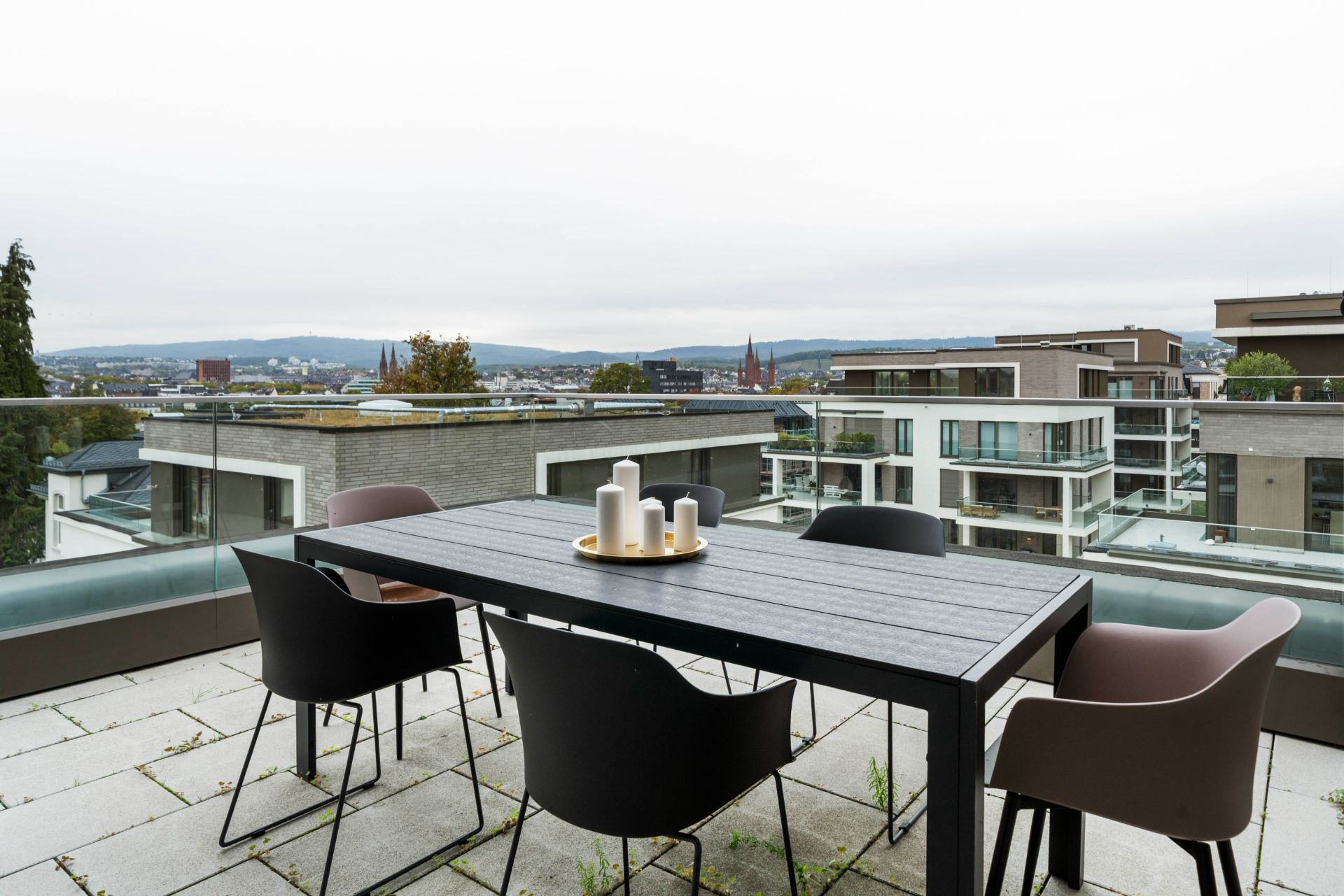 Moderne Terrasse mit Tisch, fünf Stühlen und dekorativen Kerzen, Blick auf Wohnhäuser und Stadtlandschaft.