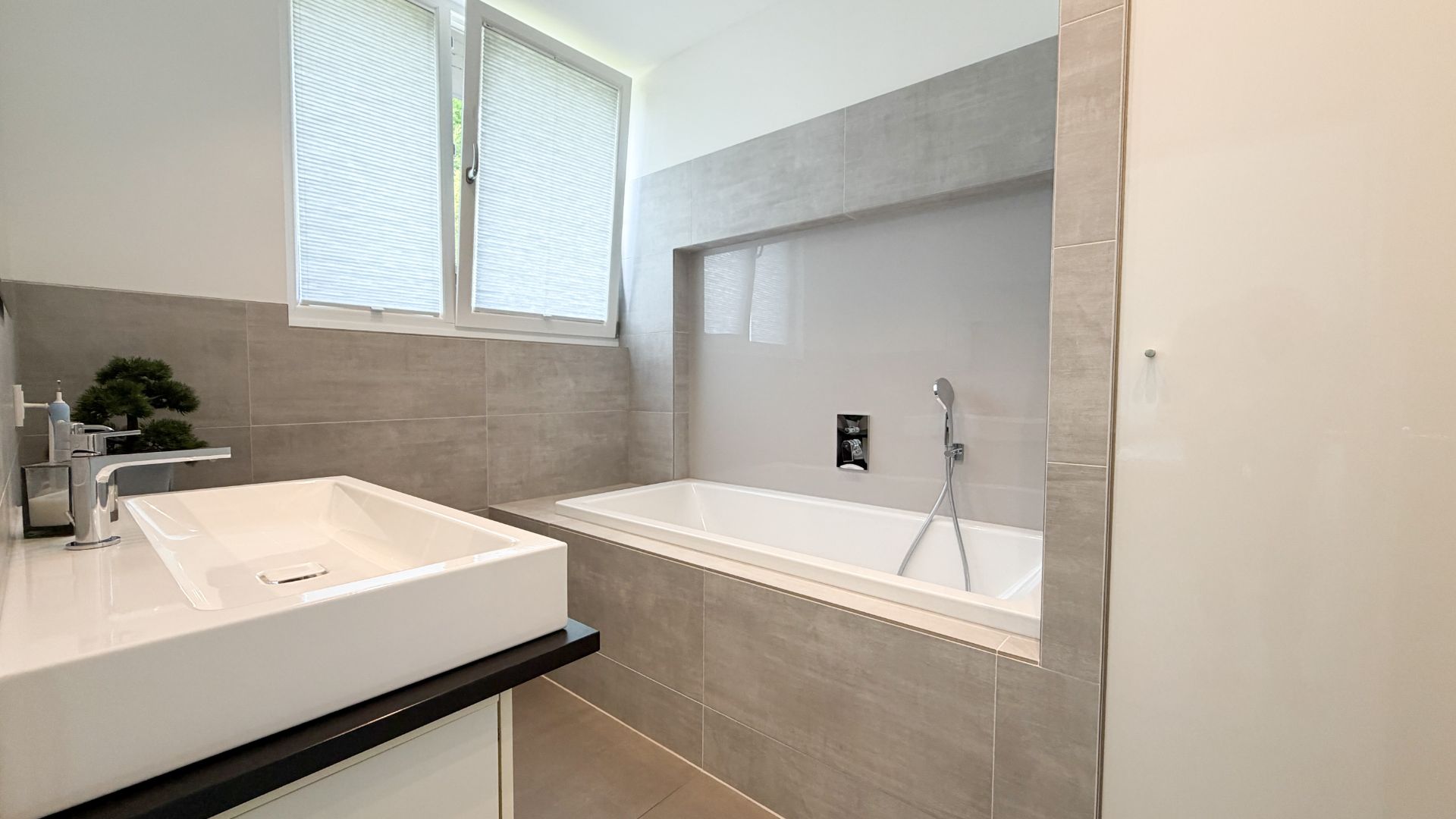 Modernes Badezimmer mit Waschbecken, Badewanne und zwei Fenstern mit Plissees.