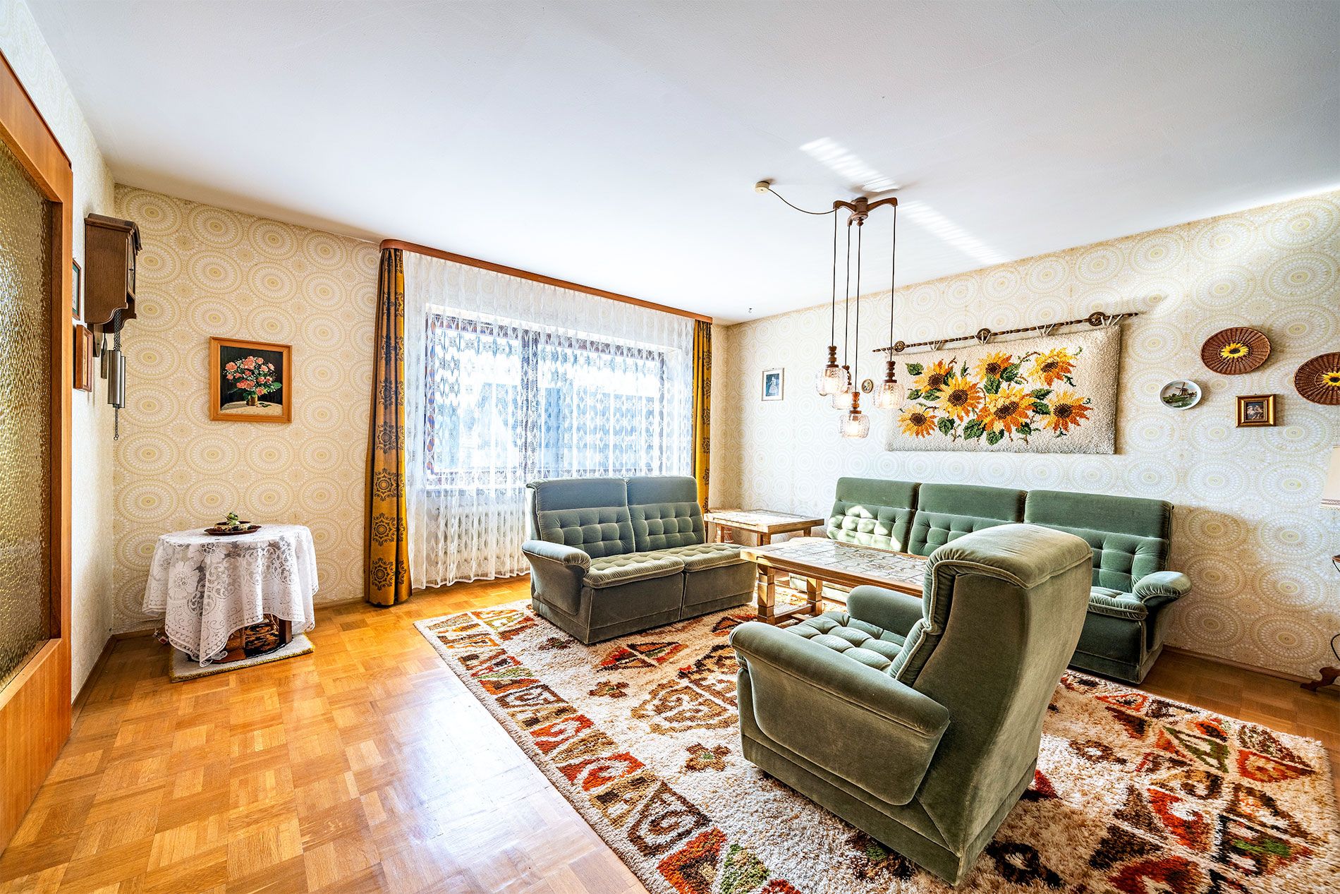Wohnzimmer mit grünen Polstermöbeln, Teppich, Couchtisch und Wanddekoration, großes Fenster mit Gardinen.